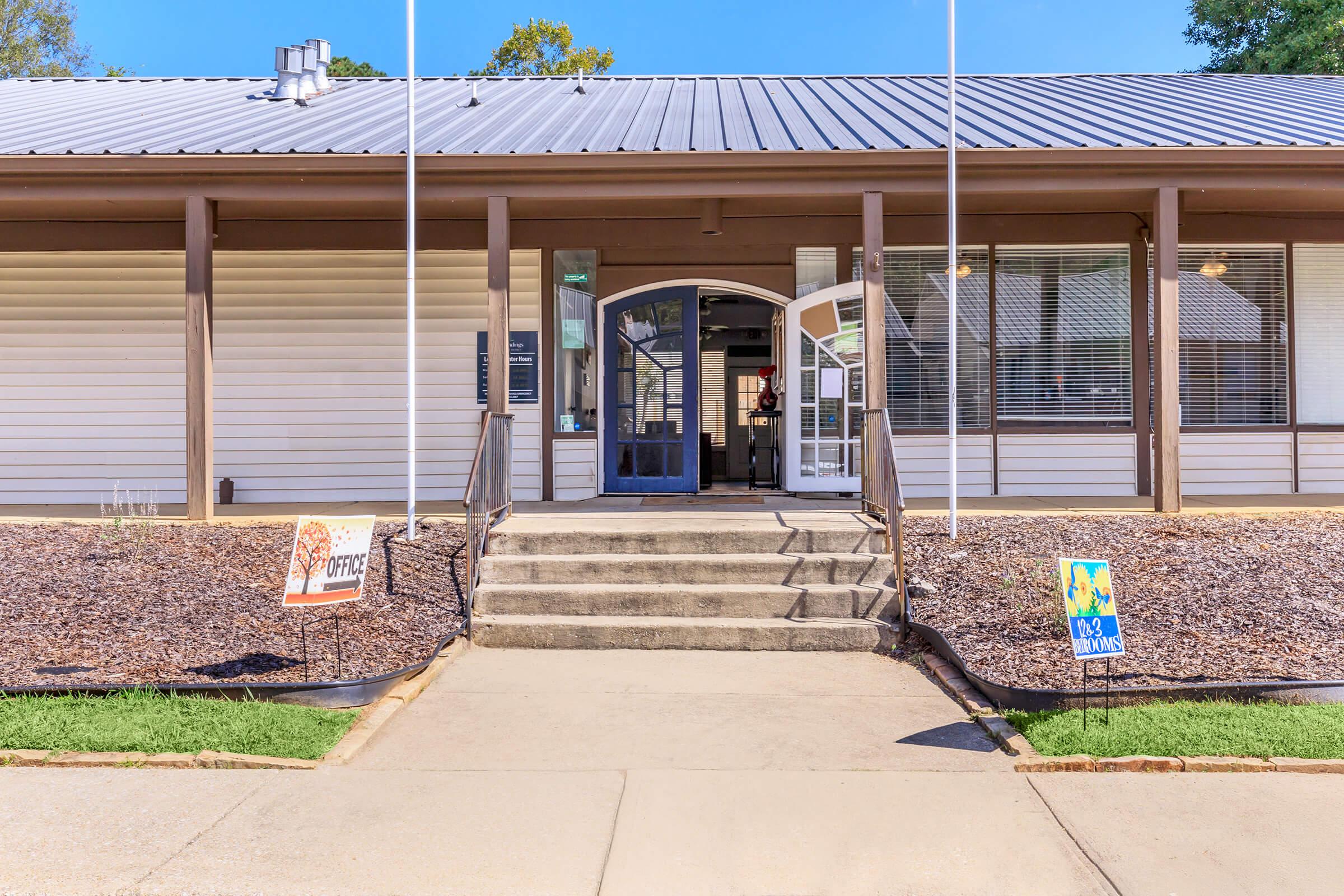 Exterior view of a single-story building with a gray metal roof, featuring a set of double doors at the entrance. There are two flags in front and decorative signs on either side of the walkway leading up to the door, surrounded by a landscaped area with mulch and grass.