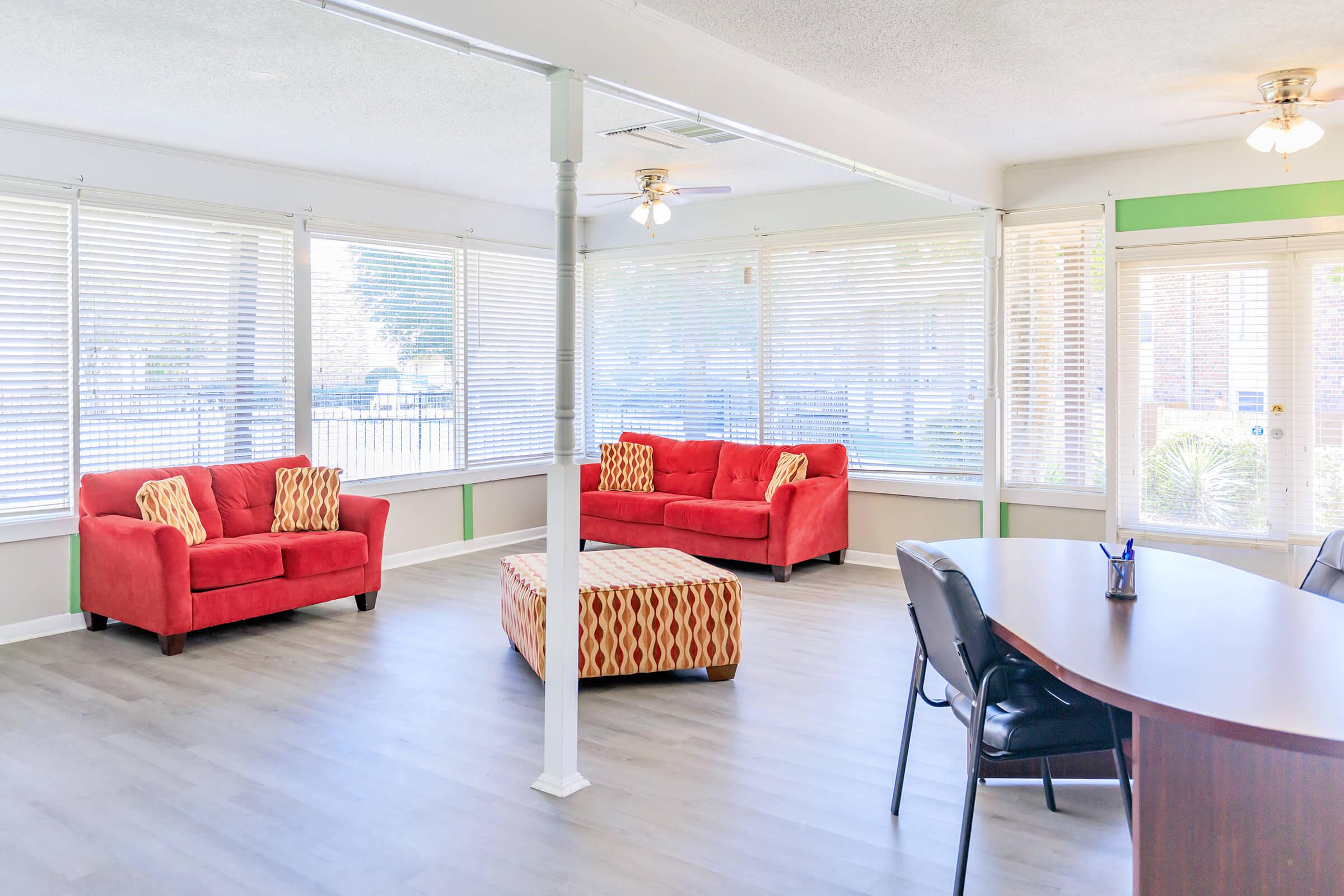 Bright and airy interior of a living room with large windows adorned with blinds. It features two red sofas with decorative cushions, an orange patterned ottoman, and a wooden desk with black chairs. Sunlight streams in, creating a welcoming atmosphere.