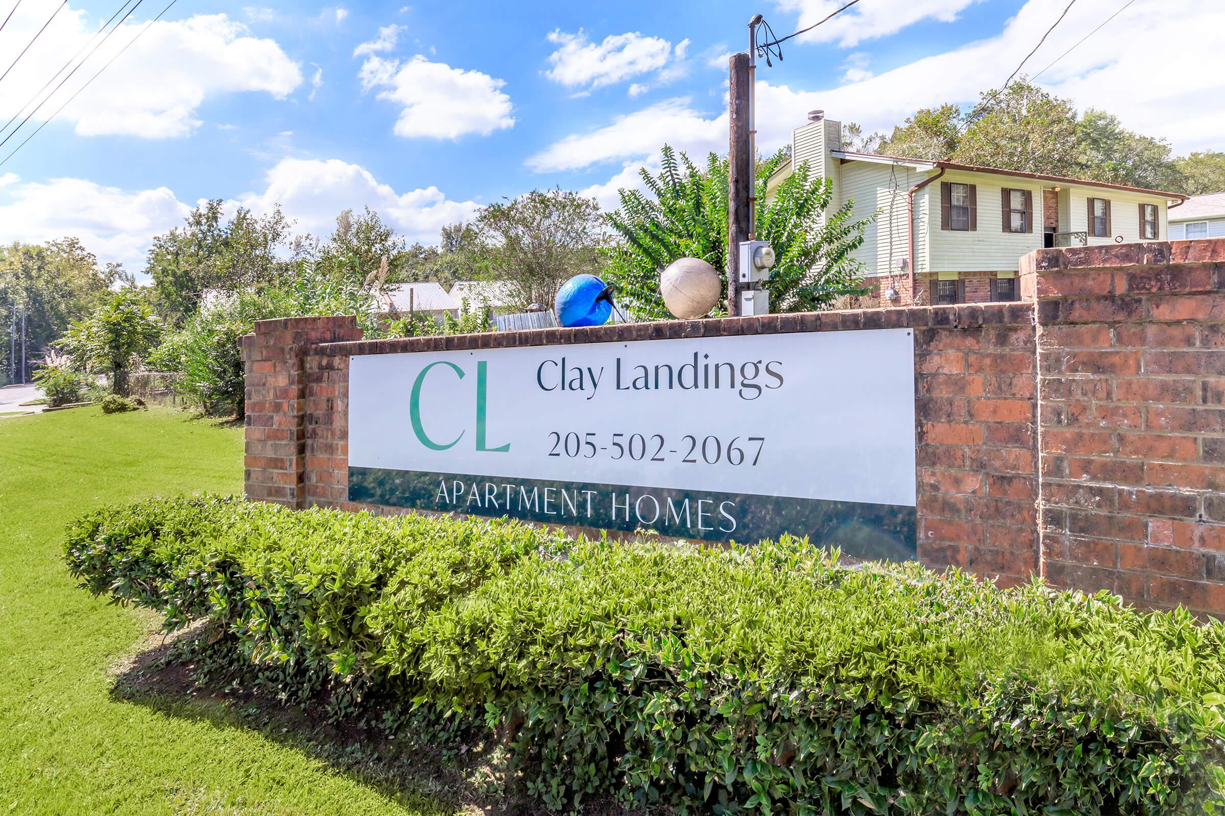 Sign for Clay Landings Apartment Homes featuring the initials "CL" and the phone number 205-502-2067, surrounded by green landscaping and a clear blue sky in the background. The sign is made of brick and is positioned near the road.