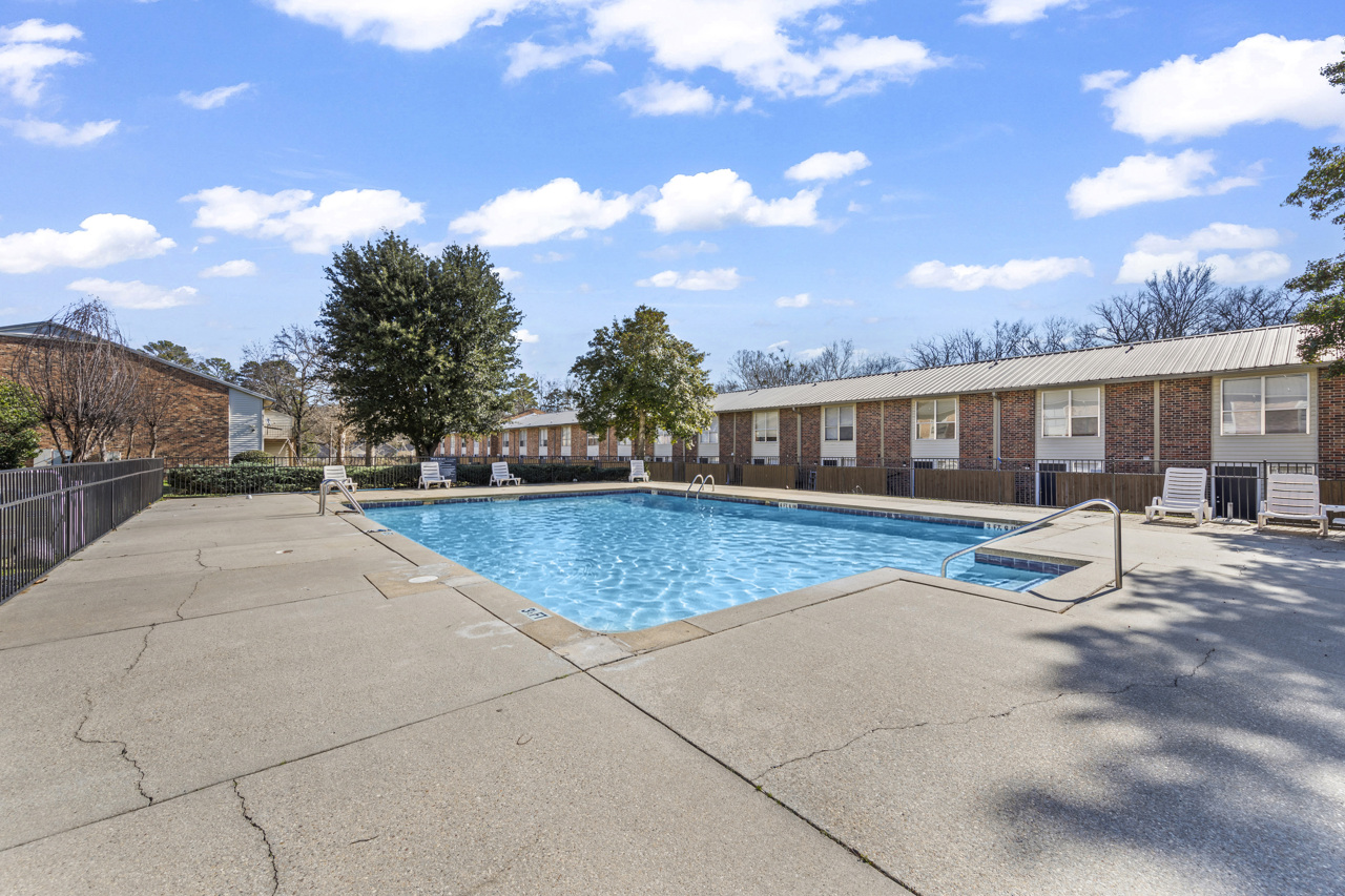 A clear, blue swimming pool surrounded by concrete, with lounge chairs on one side. In the background, there are several low brick apartment buildings and trees, all under a bright blue sky with a few clouds. The area appears well-maintained and inviting for relaxation.