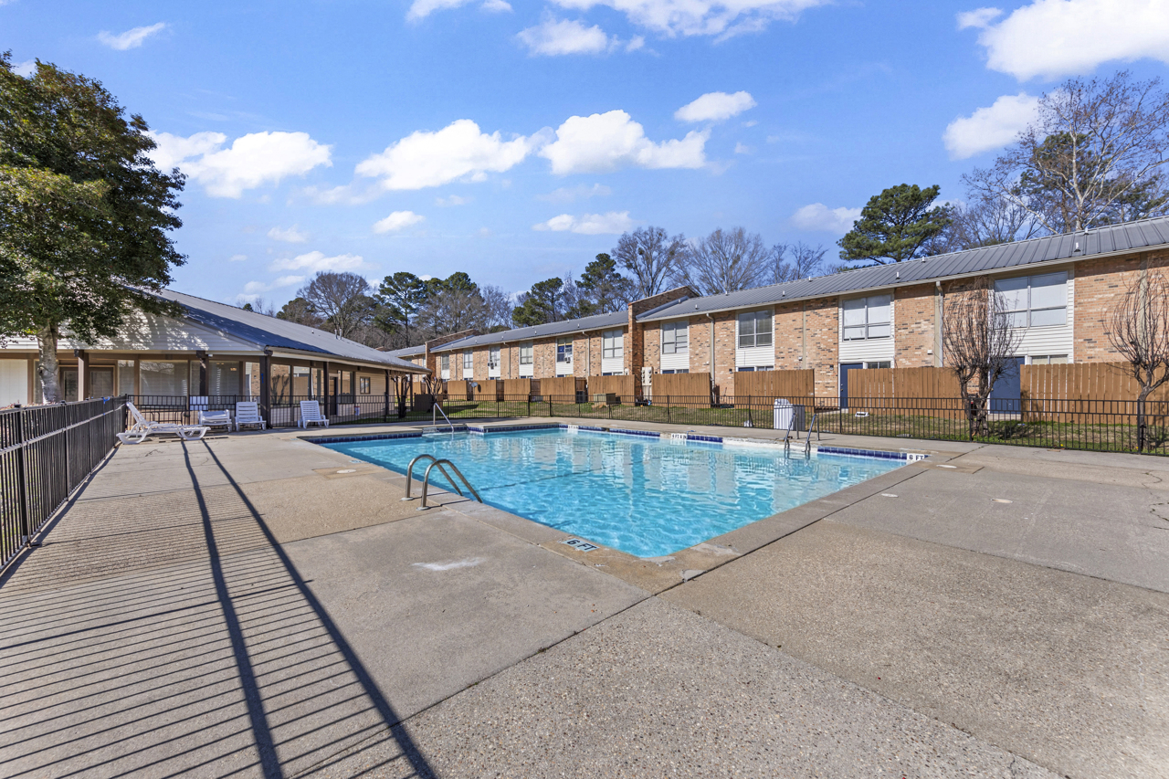 A clear swimming pool surrounded by a fenced area, with lounge chairs nearby. In the background, two brick buildings are visible, along with trees and a blue sky with scattered clouds. The pool area appears well-maintained and is likely part of a residential complex.