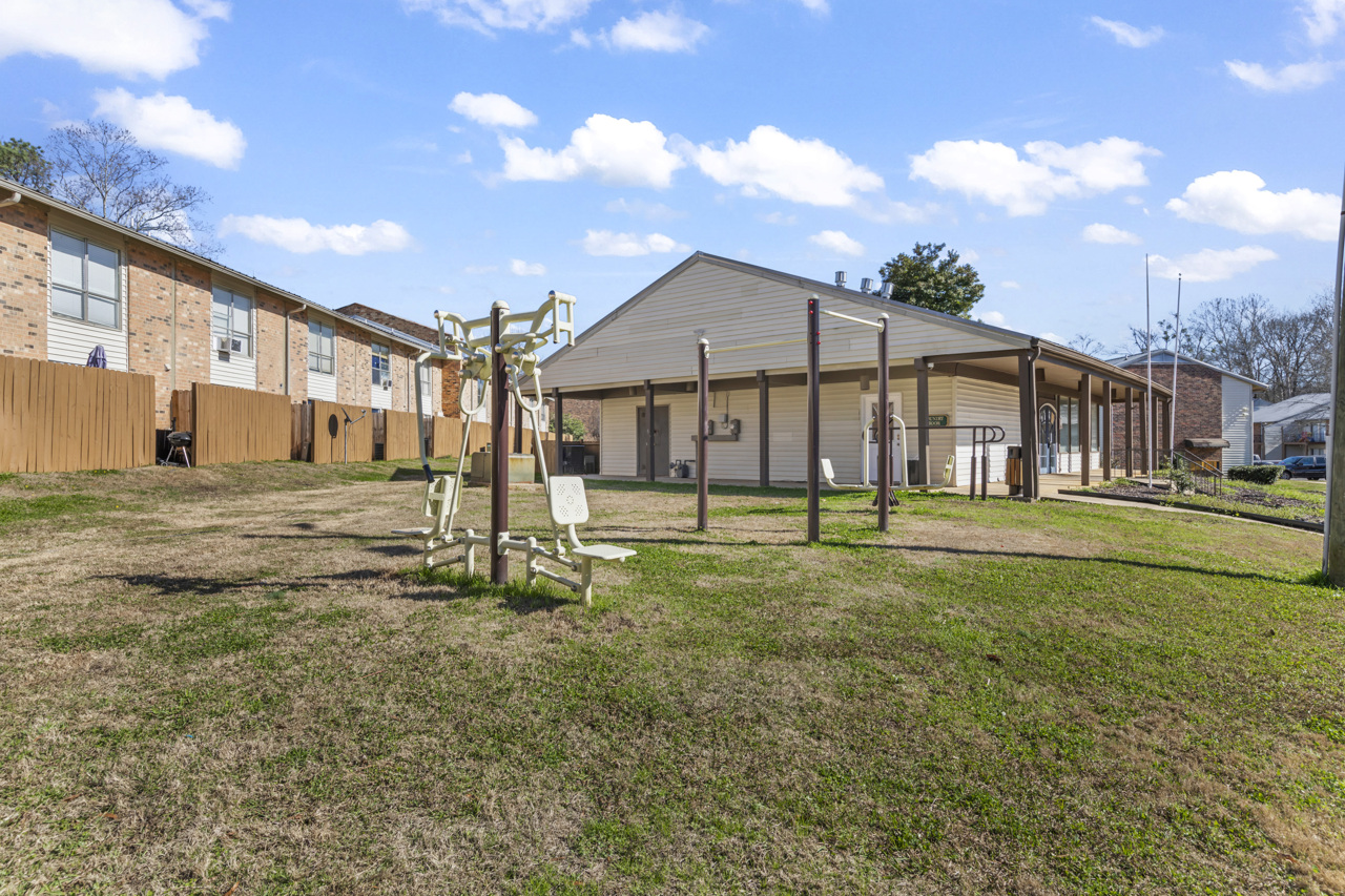 A grassy area featuring exercise equipment, with several wooden structures in the background resembling townhouses. The scene is bright with a clear blue sky and fluffy white clouds, creating a welcoming outdoor space for fitness and leisure.