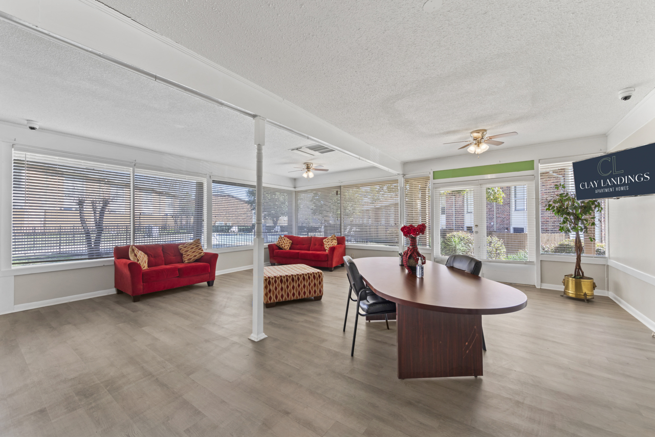 Bright, spacious common area featuring a large table with chairs, red sofas, and a decorative ottoman. Large windows allow natural light, showcasing the room's modern design and inviting atmosphere. A logo for Clay Landings is displayed in the corner.
