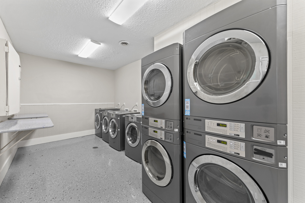 A laundry room featuring multiple stacked gray washing machines and dryers, arranged neatly against a light-colored wall. The floor is smooth and gray with a few fluorescent lights overhead, providing bright, even lighting in the space. A folding counter is visible to the left.
