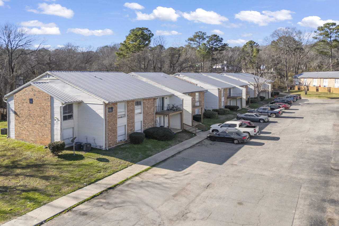 A row of apartment buildings with a mix of brick and siding exteriors. The parking area in front is partially filled with cars, and trees are visible in the background. The scene is set under a clear blue sky with a few clouds.