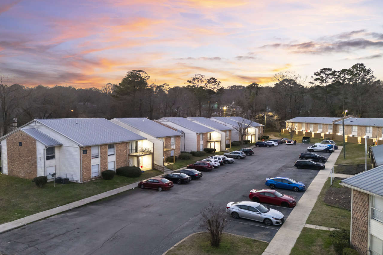 Aerial view of a residential apartment complex at sunset. The scene features several buildings arranged in a row, with a parking lot filled with various cars. The sky showcases vibrant colors of orange, pink, and purple, creating a picturesque backdrop to the neighborhood.