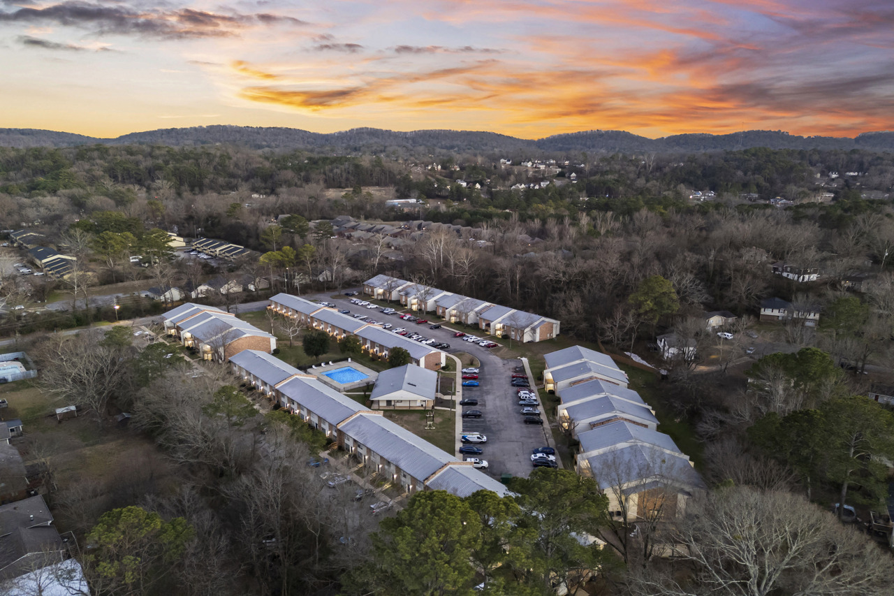 Aerial view of a residential complex featuring multiple buildings with flat roofs, parking areas filled with cars, a swimming pool in the center, and surrounded by trees. In the background, hills and a colorful sunset sky can be seen, creating a tranquil atmosphere.