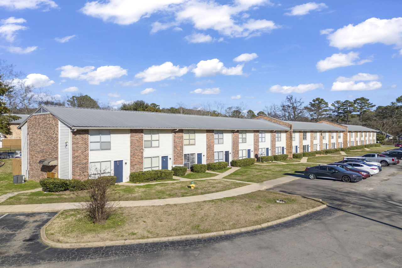 A view of a row of brick apartment buildings with a gray metal roof, surrounded by grassy areas and small shrubs. The parking lot features several parked cars, and the sky is partly cloudy, creating a bright and inviting atmosphere.