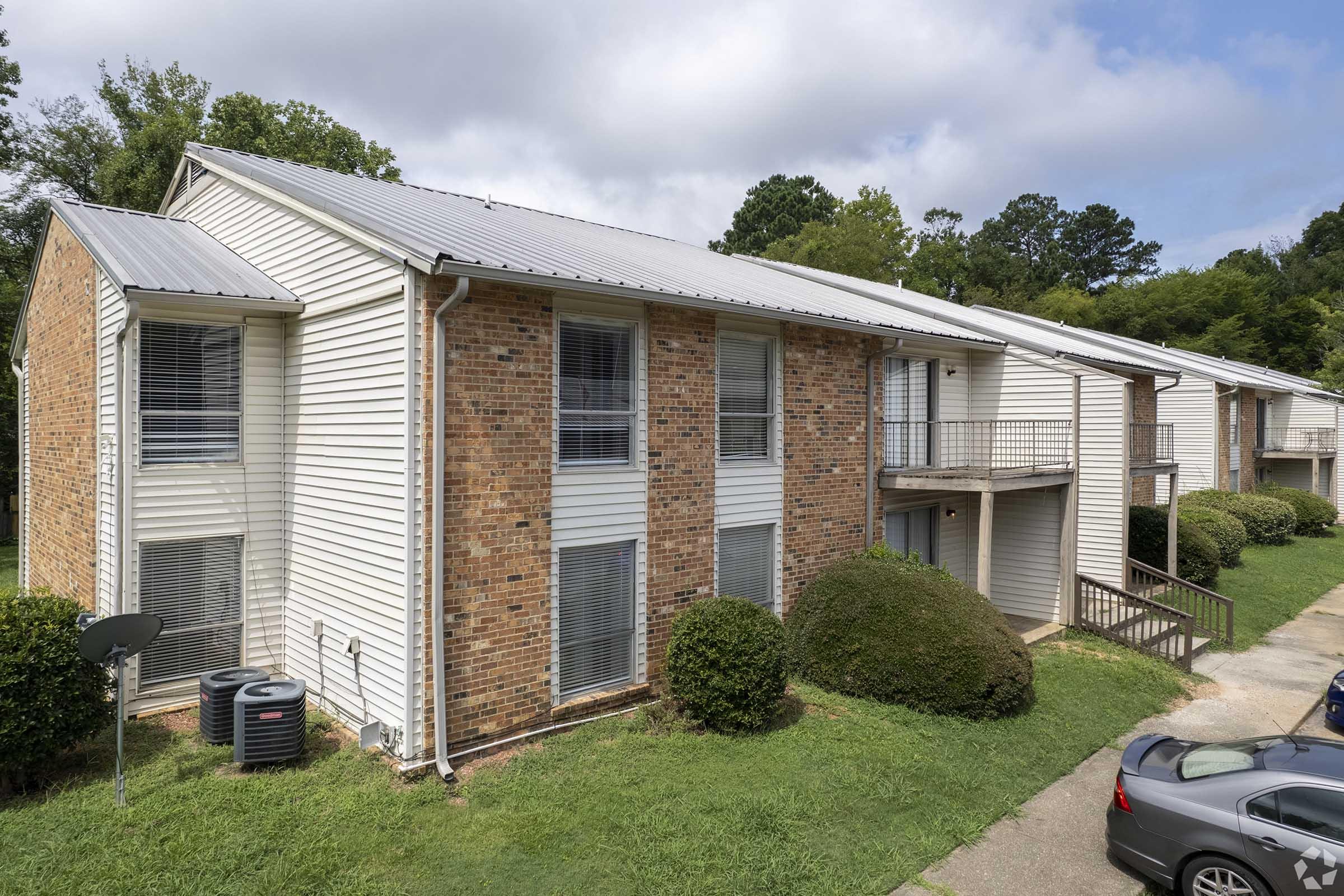 A view of a two-story apartment building made of brick and siding, featuring multiple units with balconies. The surrounding area has well-maintained grass and shrubs, along with parked cars and a satellite dish. The sky is partly cloudy, adding to the residential atmosphere.