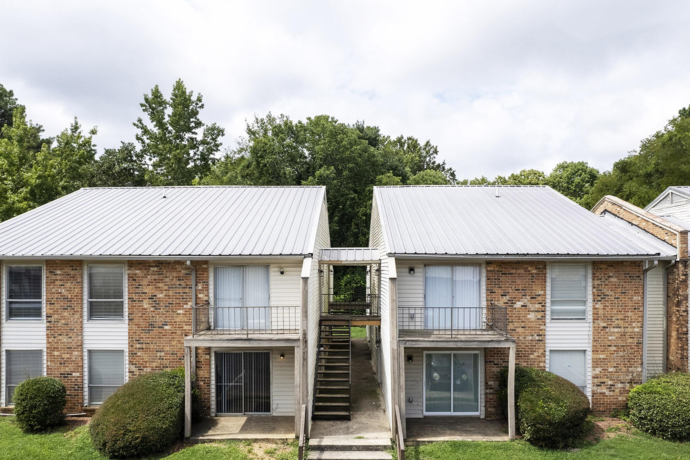 A view of a two-story brick apartment building with a metal roof. Each side features a set of stairs leading to individual balconies. The surrounding area includes green trees and shrubs, creating a natural setting. The sky is partially cloudy.