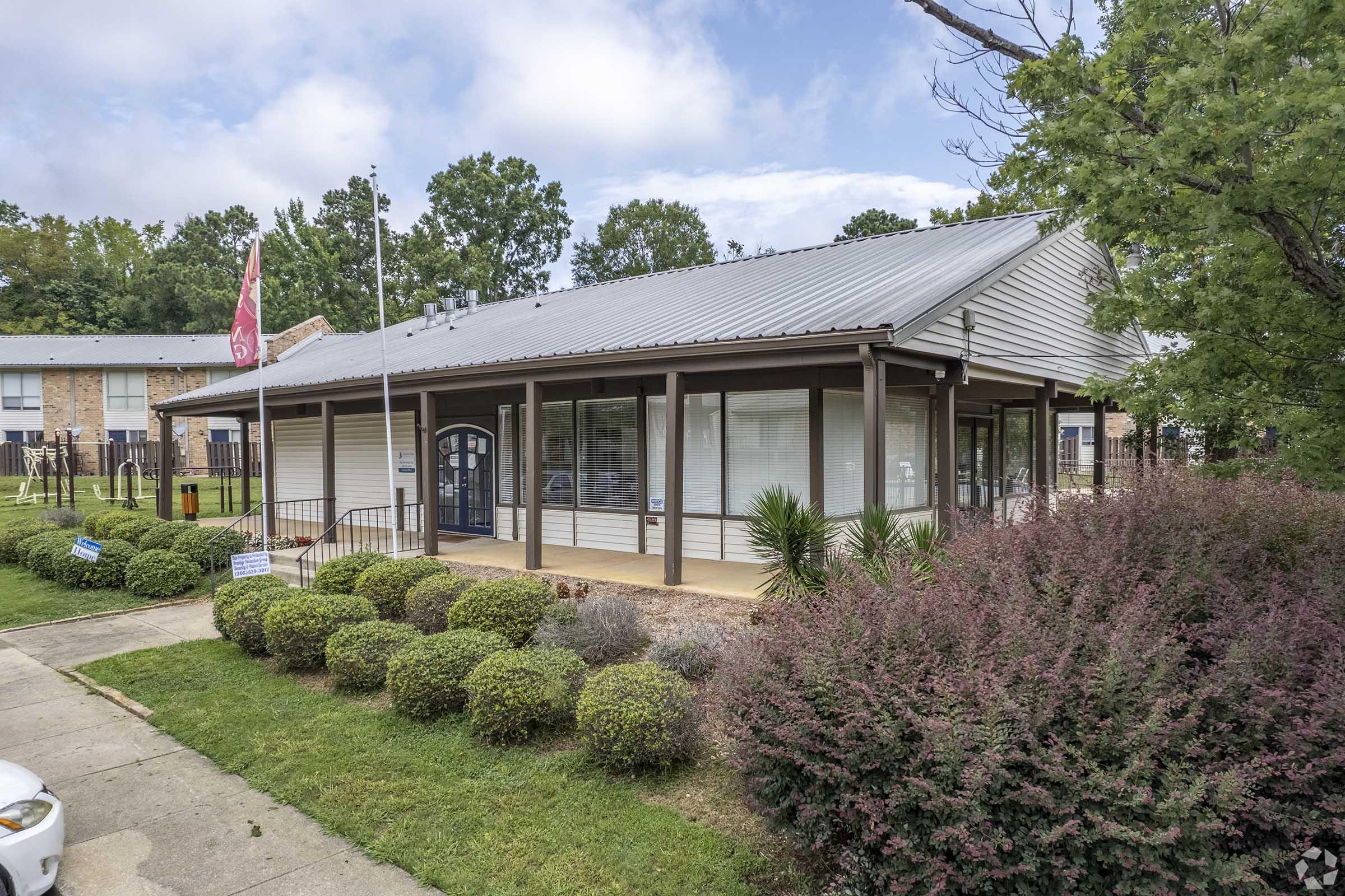 A single-story building with a metal roof, featuring large windows and a welcoming entrance. The front has landscaped greenery, including hedges and bushes, with a flagpole nearby. The background shows trees and additional structures, indicating a community setting.