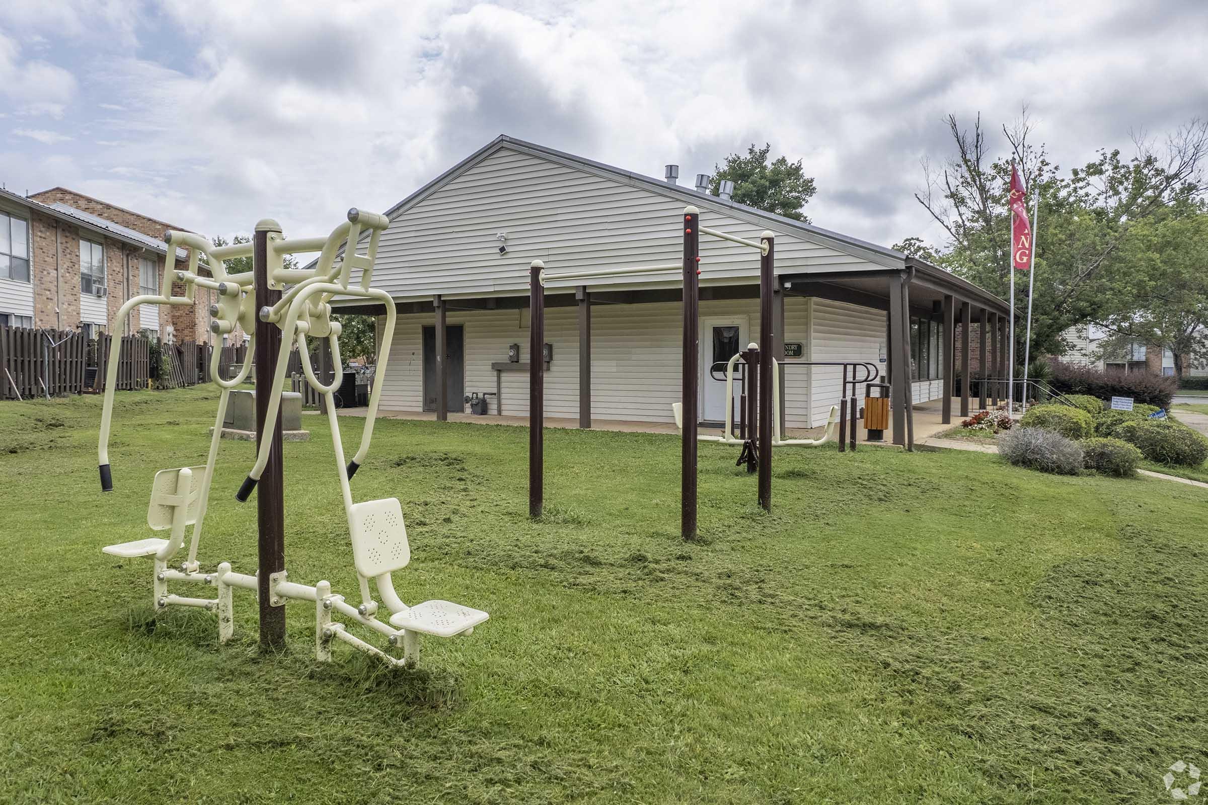 Outdoor fitness area with exercise equipment, including leg press and seated row machines, set on a green lawn. A building with a porch is in the background, and trees are visible nearby. The scene is partially cloudy, creating a bright and inviting atmosphere.