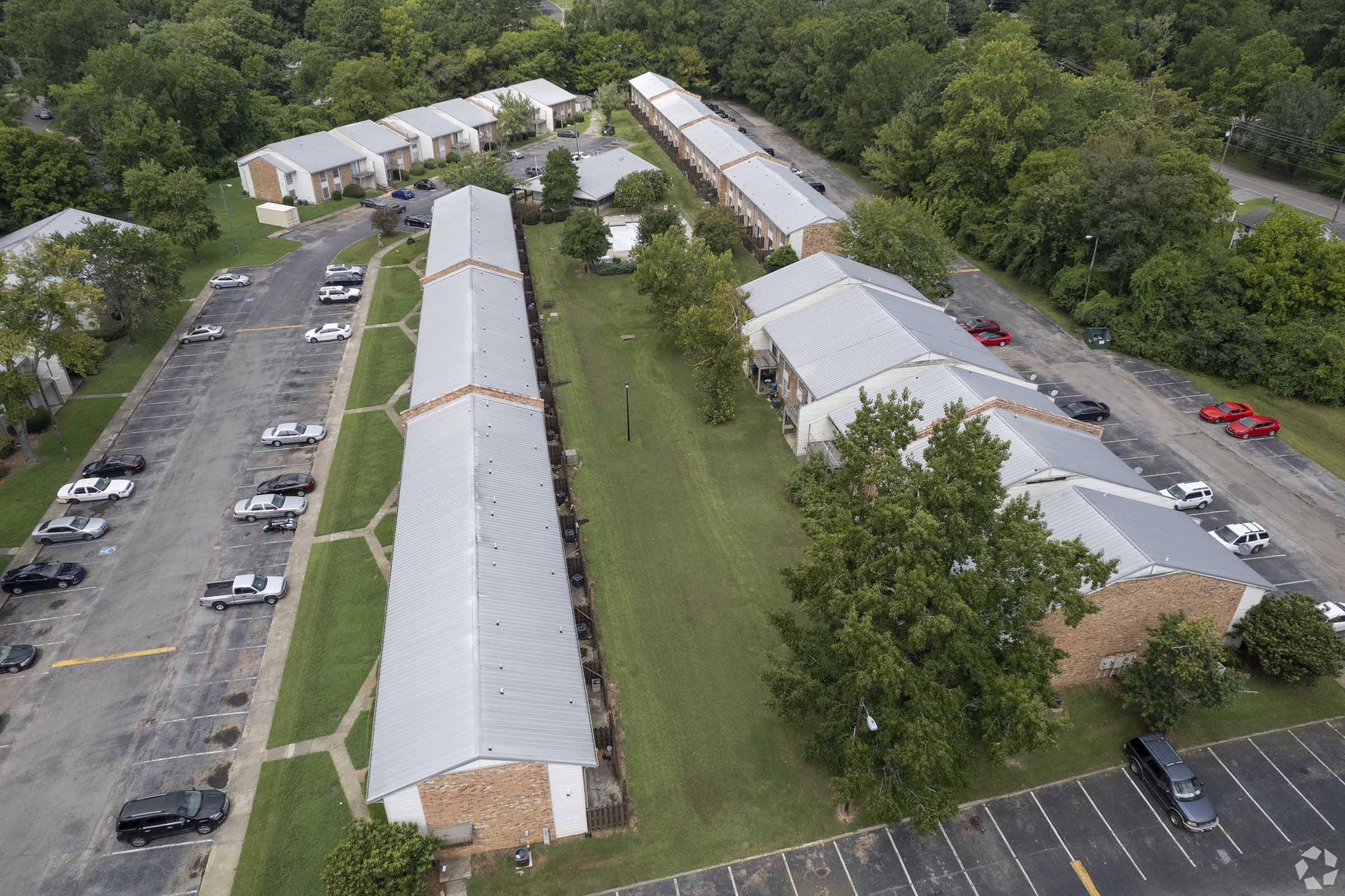 Aerial view of a residential complex featuring several two-story buildings with metal roofs, surrounded by green lawns and trees. Parking lots with parked cars are visible along the pathways connecting the buildings, indicating a well-maintained neighborhood layout.