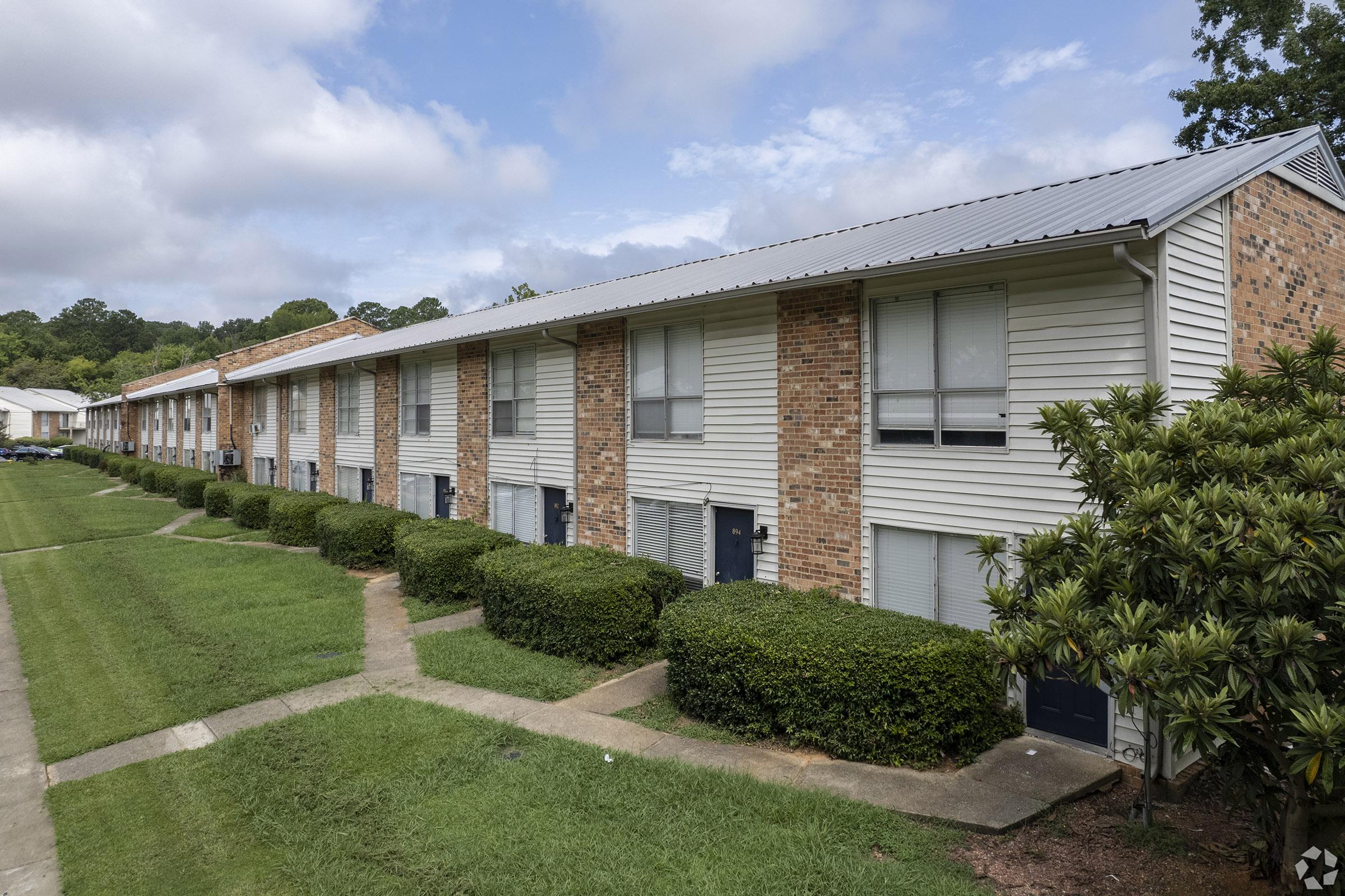 A row of two-story apartment buildings with brick and siding exteriors, surrounded by well-maintained lawns and hedges. The sky is partly cloudy, suggesting a pleasant day. The pathway in front of the apartments leads to individual entrances.