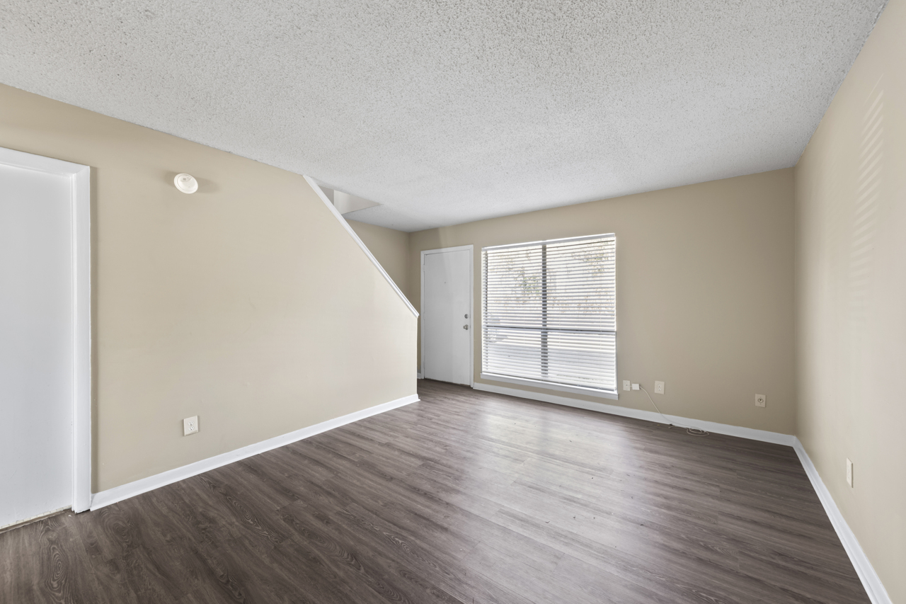 A spacious, empty living room with light brown walls and dark wood-like flooring. Large windows with blinds allow natural light, and there is a door leading outside. A staircase is visible in the background, and a white door is on the left side of the image. The room has a clean and modern feel.