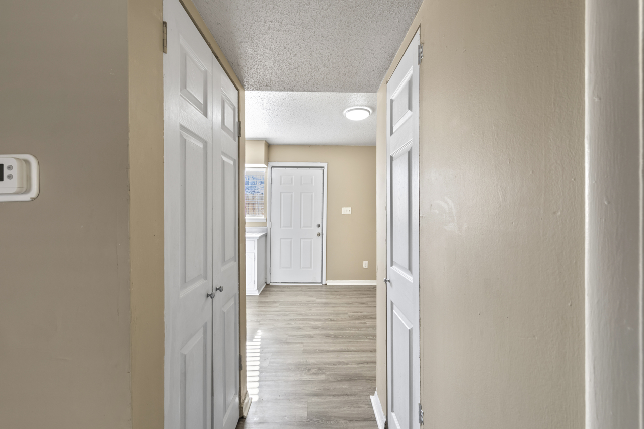 View of a hallway leading to a bright, open space. Two closed white doors are visible on the left, and a white door at the end leads to another room. The walls are painted a warm beige color, and the floor features light-colored laminate flooring. Natural light enters from a window, creating a welcoming atmosphere.