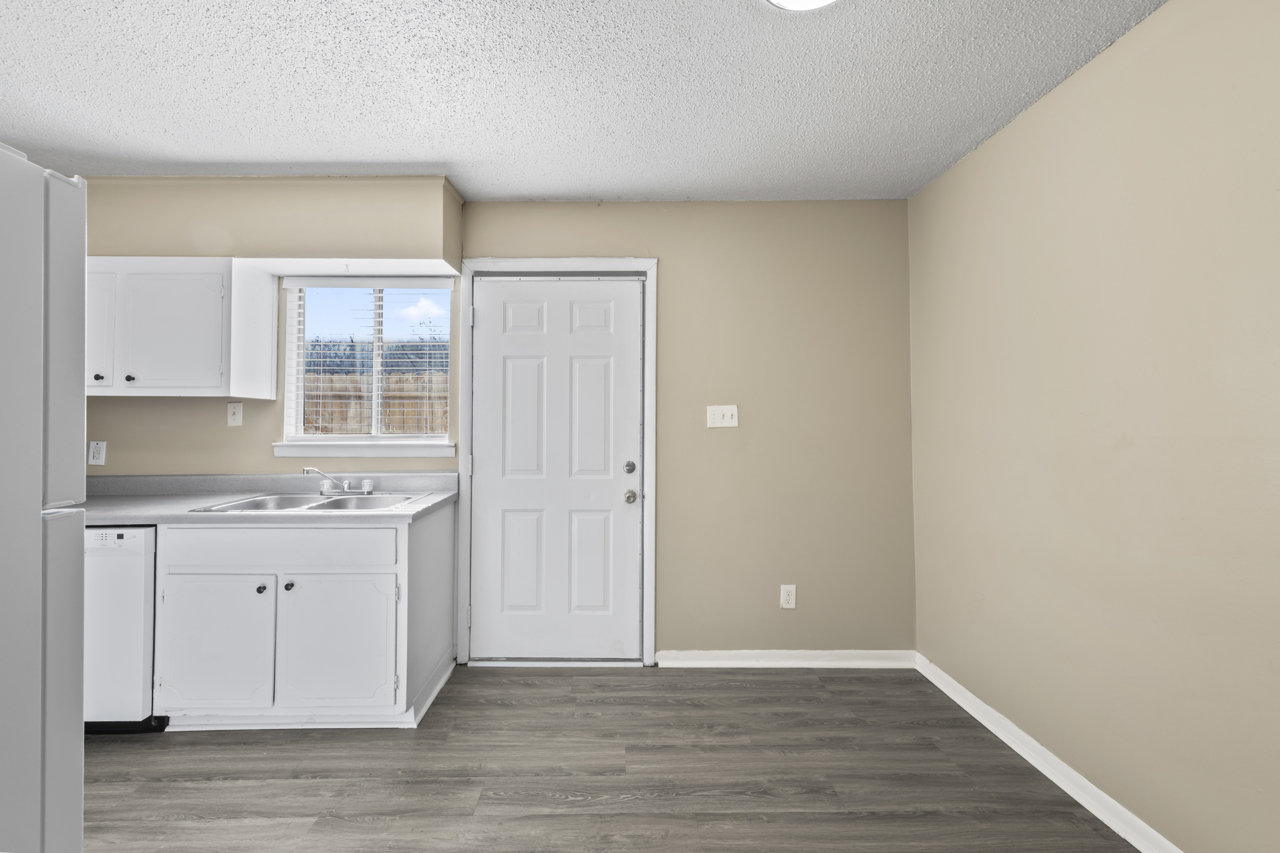 A simple kitchen with white cabinets and appliances, a sink, and a door leading outside. The walls are painted a light beige, and there is a window allowing natural light to enter. The floor is a light-colored laminate. The overall space is clean and uncluttered.