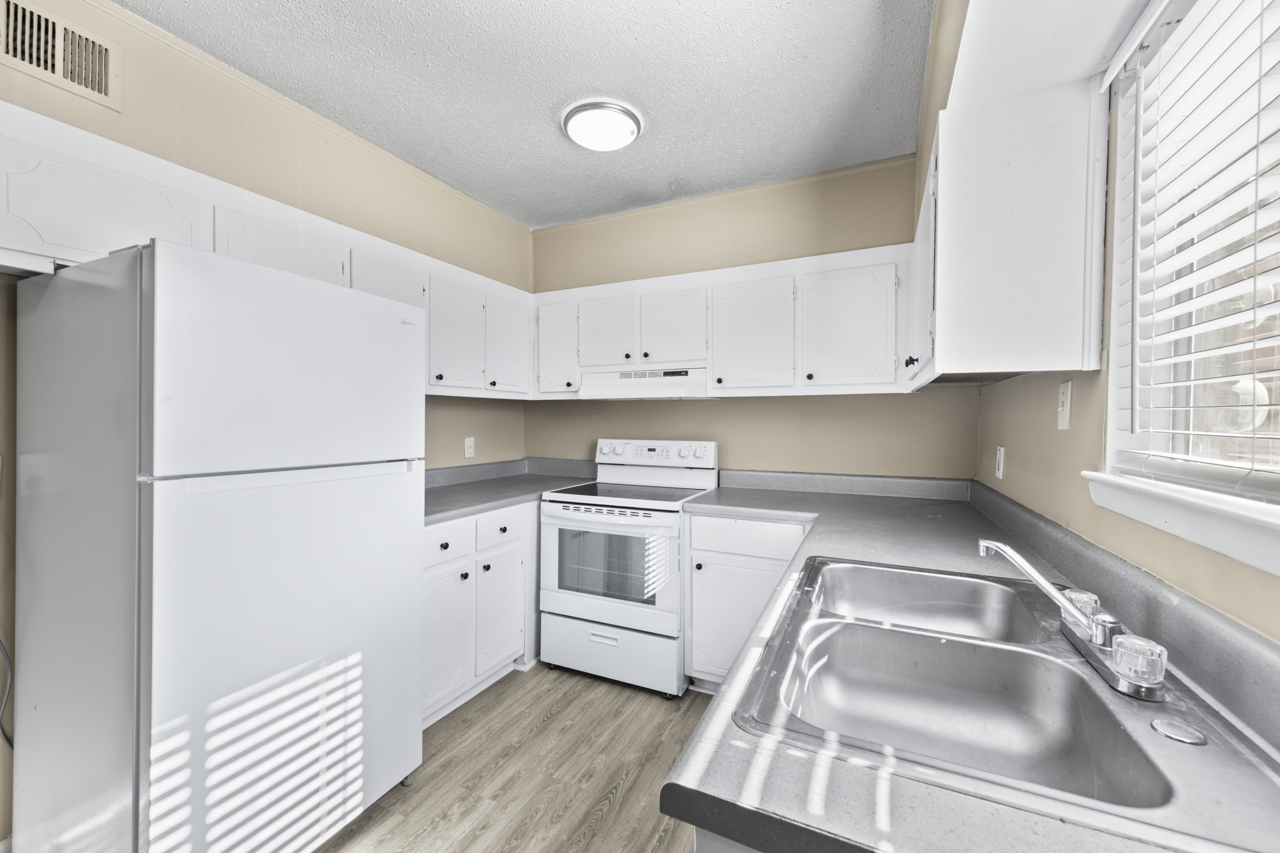 A bright, modern kitchen featuring white cabinets, a white refrigerator, and a white stove with an oven. A double sink is positioned under a window that allows natural light to fill the space. The flooring is light wood, and the walls are painted a soft beige. The overall atmosphere is clean and inviting.
