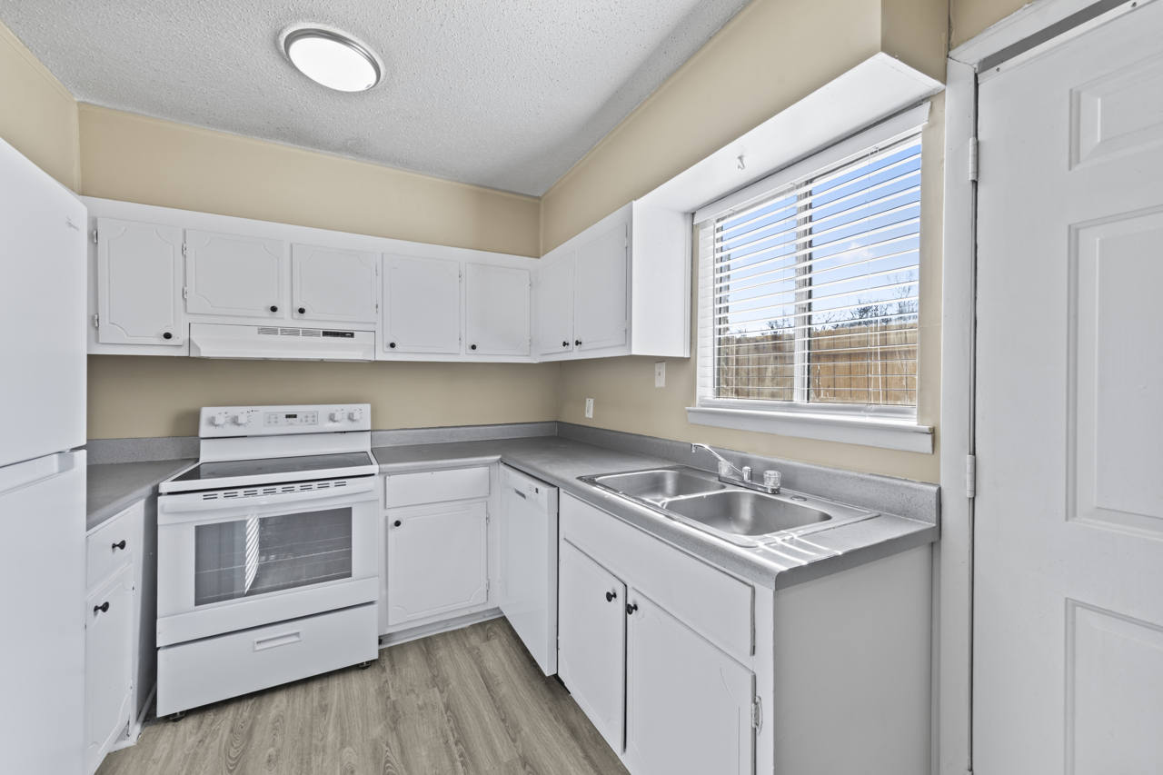 A bright, modern kitchen featuring white cabinets, a stainless steel oven, and a double sink. The space includes a window with blinds that allows natural light, and light-colored flooring complements the neutral walls. The room is uncluttered and has a clean, inviting appearance.
