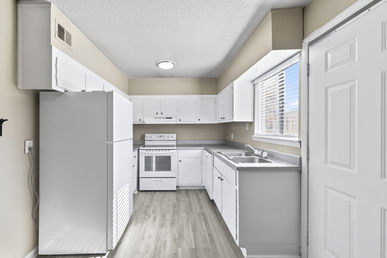 A bright and clean kitchen with white cabinets and appliances. Features a refrigerator, stove, and double sink, along with a window allowing natural light to illuminate the space. The flooring is a light-colored laminate, creating a spacious and modern feel.