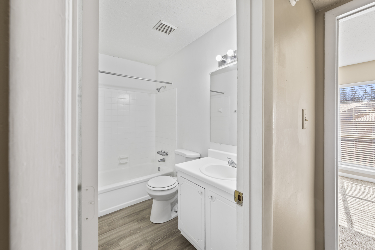 A clean, modern bathroom featuring a bathtub with a shower, a toilet, and a sink with a mirror above. The walls are painted white, and the flooring is light wood. Natural light streams in from a window, enhancing the spacious, bright appearance of the room.