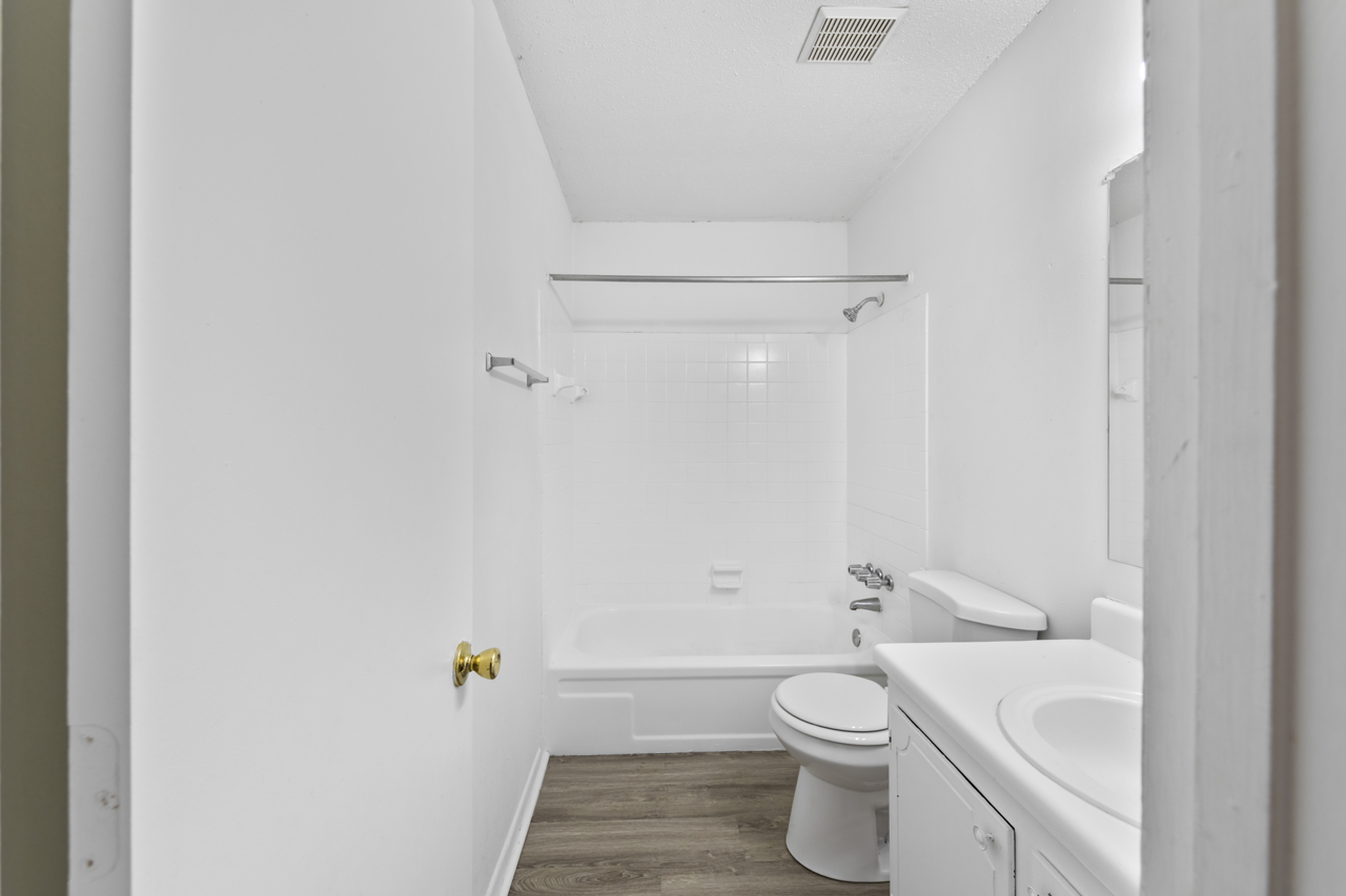 A simple, modern bathroom featuring a bathtub with a shower, a toilet, and a sink with a countertop. The walls are white, and the floor has a wooden texture. The space is well-lit and uncluttered. A mirror is mounted above the sink, and there is a small ventilation fan on the ceiling.