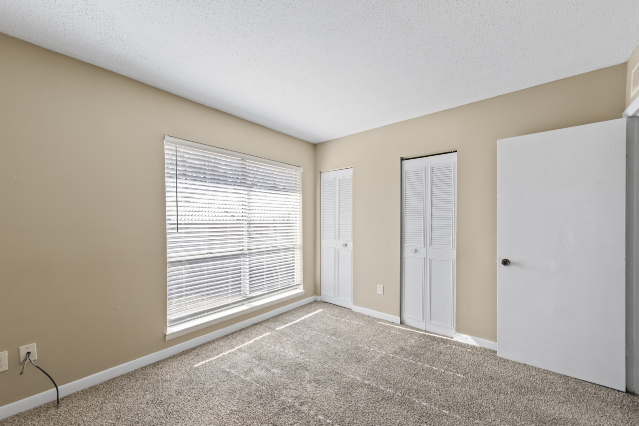 A well-lit room with beige walls and carpeted flooring. There is a large window with blinds providing natural light, a closet with double doors on one side, and a door leading to another space on the right. The room is empty, creating a spacious and airy feel.