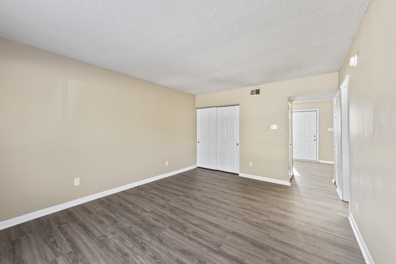 A spacious, empty room with light beige walls and laminate flooring. The room features a large closet with white double doors on one side and an open doorway leading to another room on the other side. Natural light is entering through the windows, creating a bright atmosphere.