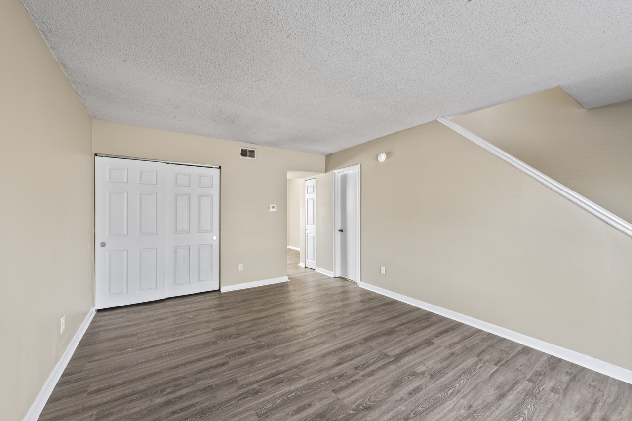 An empty room with light beige walls and wooden flooring. A closed white double-door closet is on one side, and a doorway leads to another room in the background. The space is well-lit with natural light from the windows. A staircase is visible in the corner, indicating a multi-level layout.