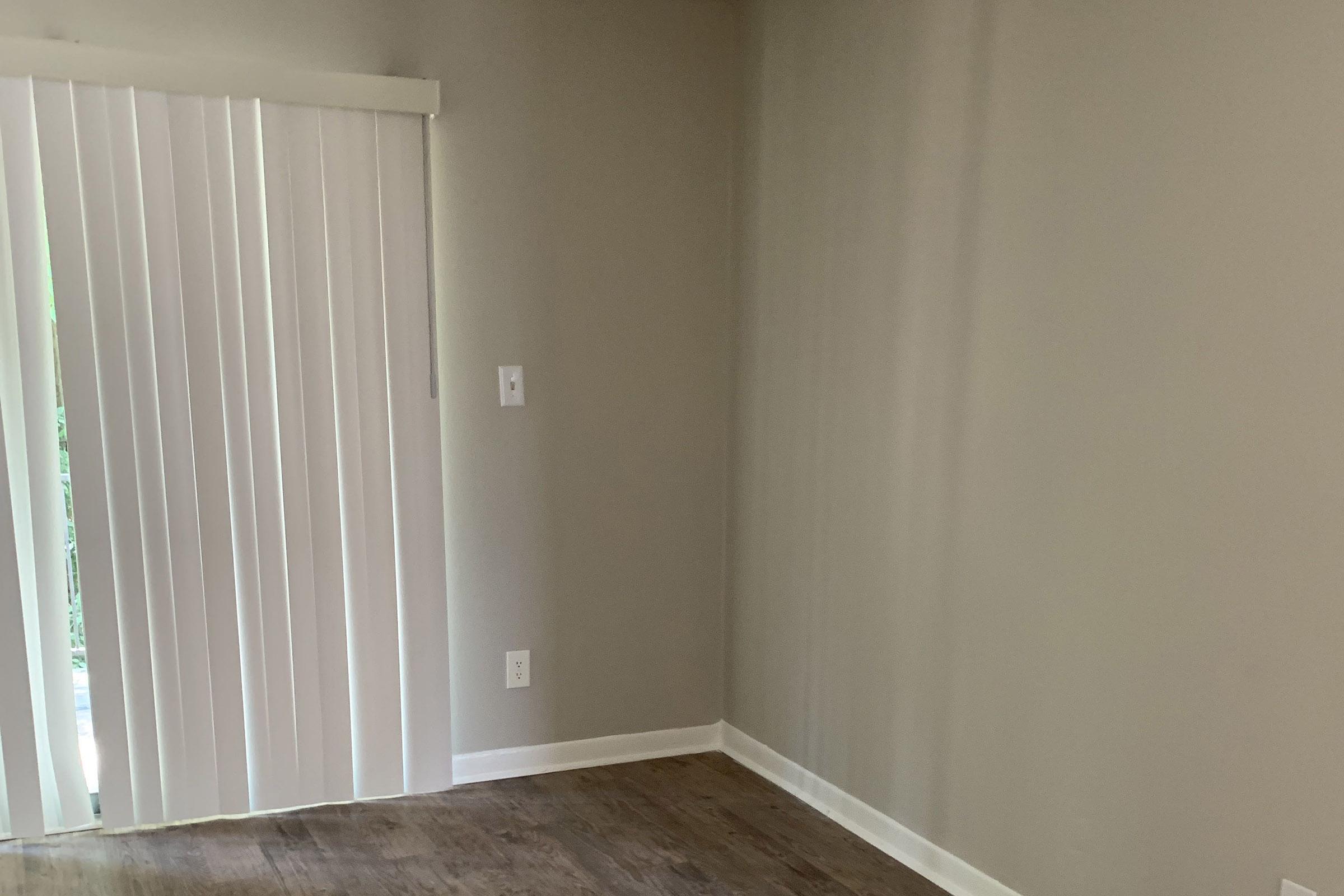 Empty room with light-colored walls and wooden flooring. A vertical blind window is partially closed, allowing some natural light to enter. No furniture or decorations are visible, creating a minimalist and spacious atmosphere.
