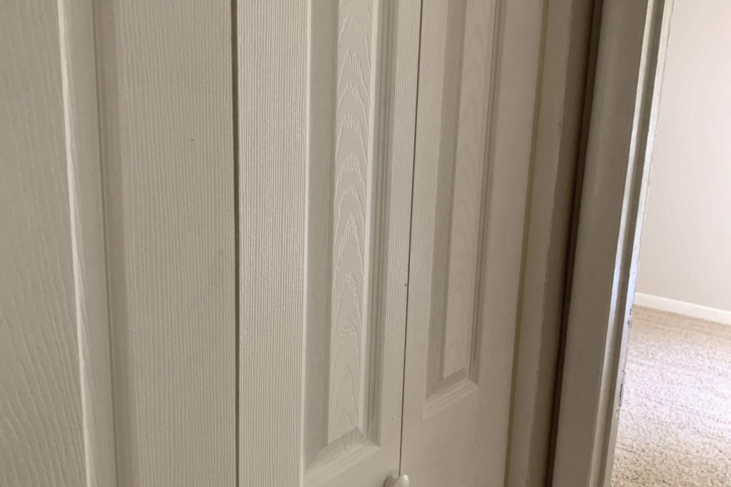 A close-up view of a white, paneled closet door with a textured surface. The door is partially open, revealing a glimpse of the interior space beyond, which appears light and neutral in color. The flooring is carpeted, adding a cozy touch to the setting.
