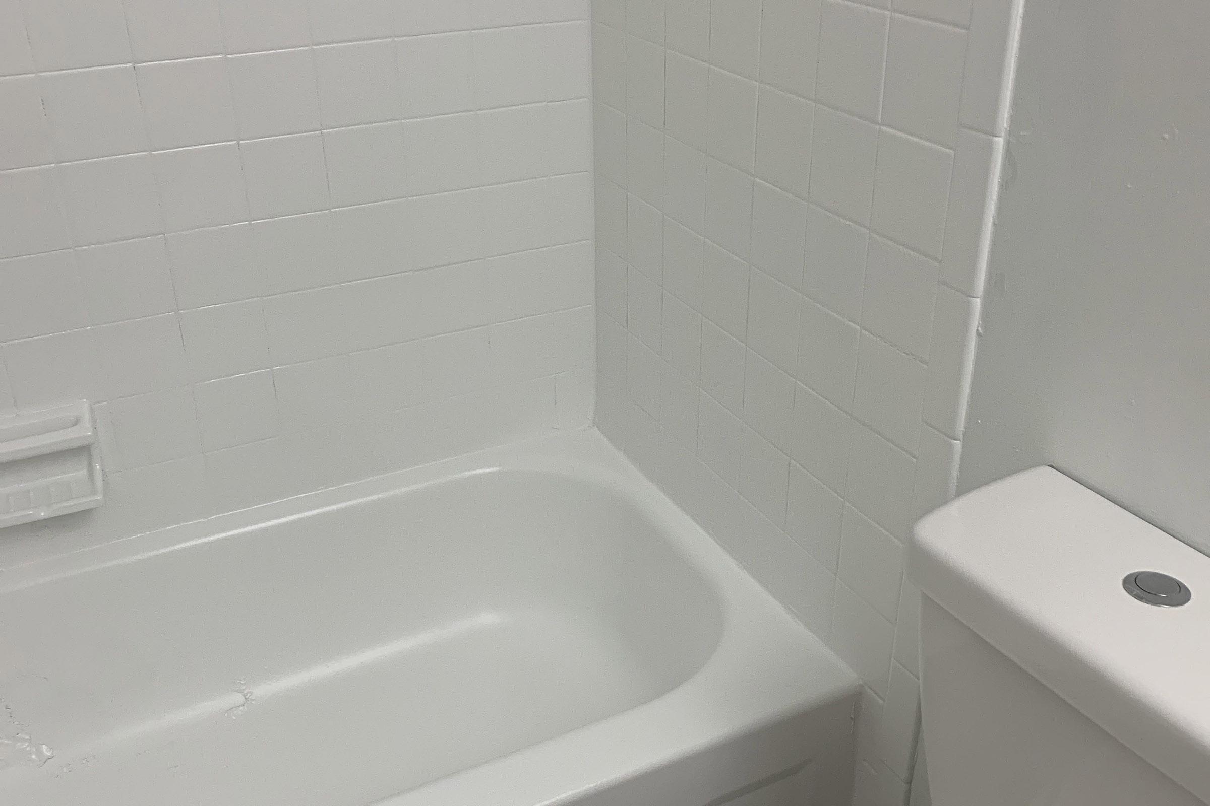 A clean, simple bathroom featuring a white bathtub and a white toilet against a tiled wall. The tiles are white and smoothly arranged, creating a minimalist aesthetic. The bathtub is empty, and the overall appearance is bright and uncluttered.