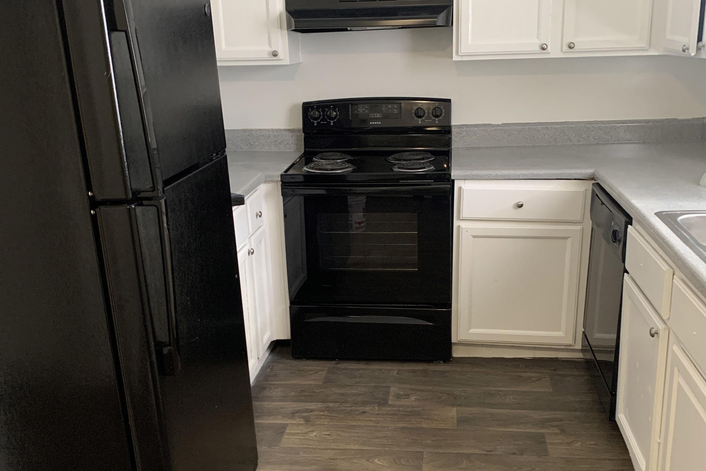 A modern kitchen featuring a black refrigerator and an electric stove. The cabinets are a mix of white and gray, with a light gray countertop. The flooring is dark wood, creating a contemporary look in the space. The sink is visible next to the stove, and the kitchen is well-lit.