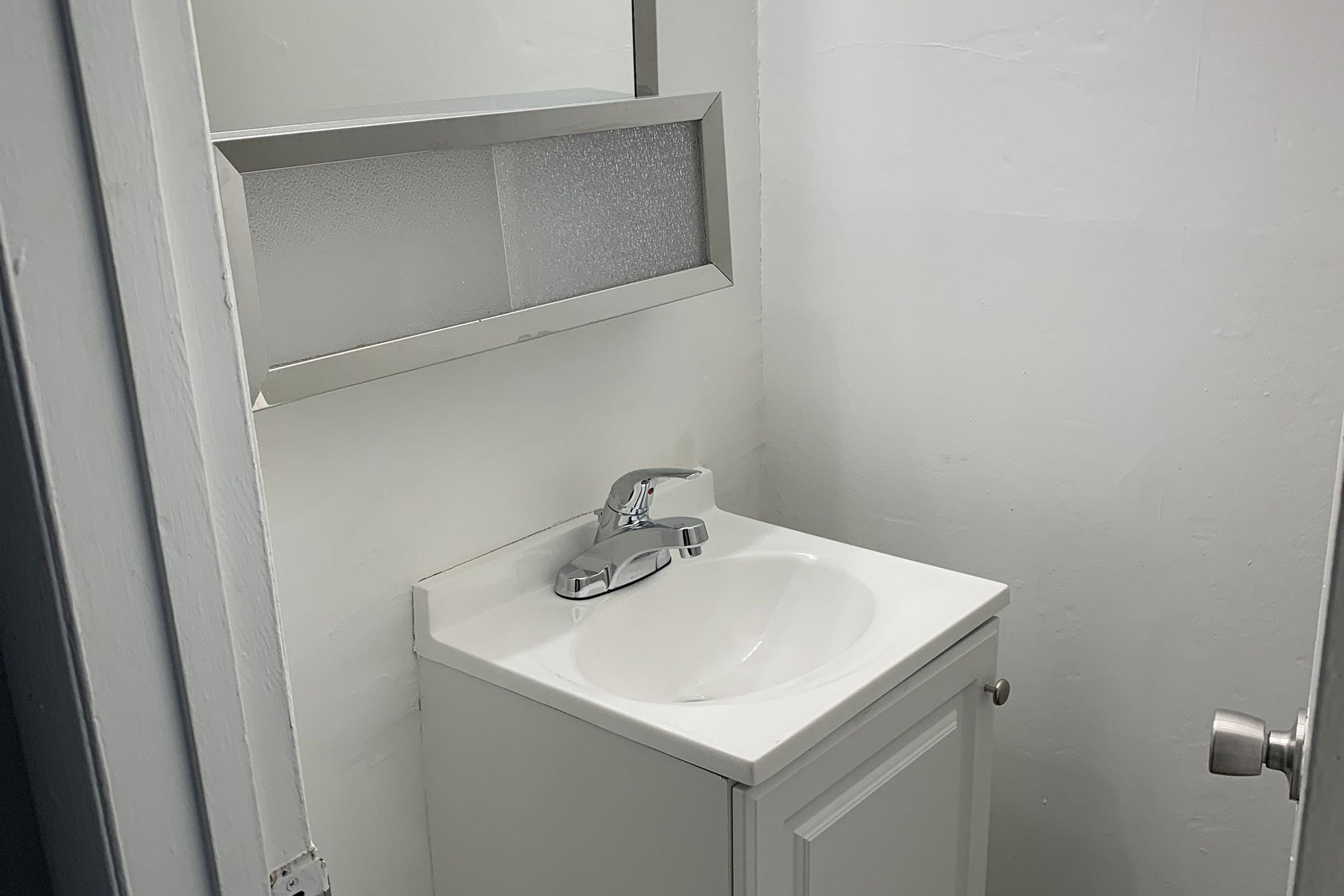 A small bathroom with a white sink and faucet mounted on a cabinet. A mirror is positioned above the sink. The walls are plain and painted white, and there is a door visible to the right. The overall space appears clean and minimalistic.