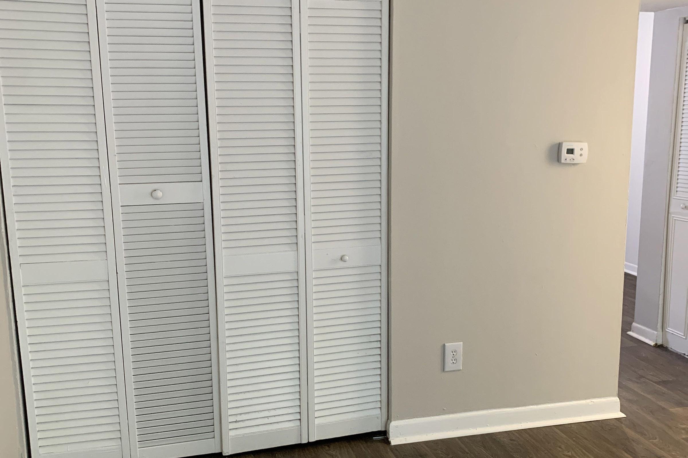 A simple room interior featuring a light-colored wall with a white sliding door closet, a small thermostat on the wall, and hardwood flooring. Part of the room is visible with neutral tones, creating a clean and minimalistic appearance.