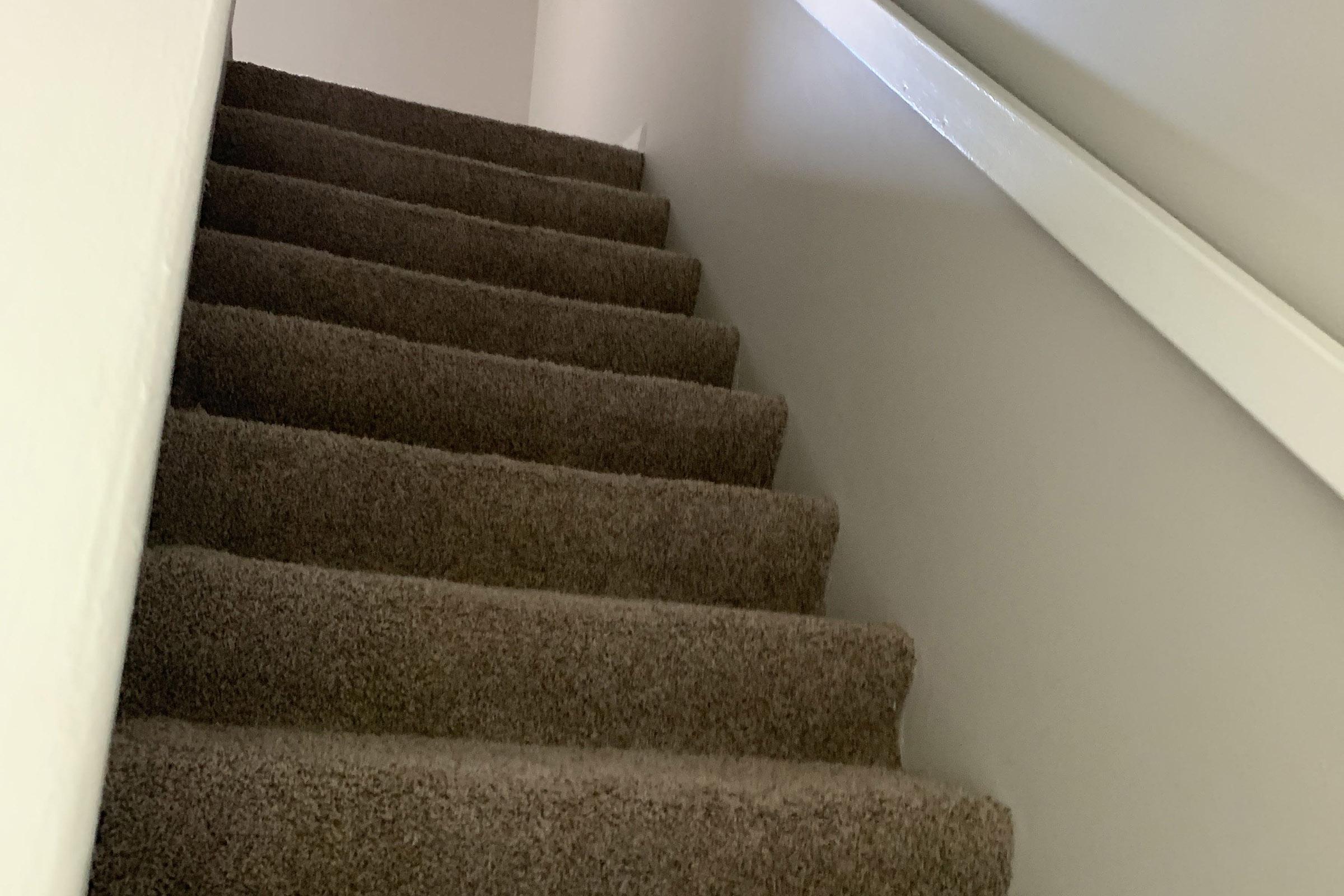 A narrow staircase with carpeted steps leading upwards, featuring a plain white wall on one side and a white railing along the top edge. The lighting is soft, creating a warm and inviting atmosphere.