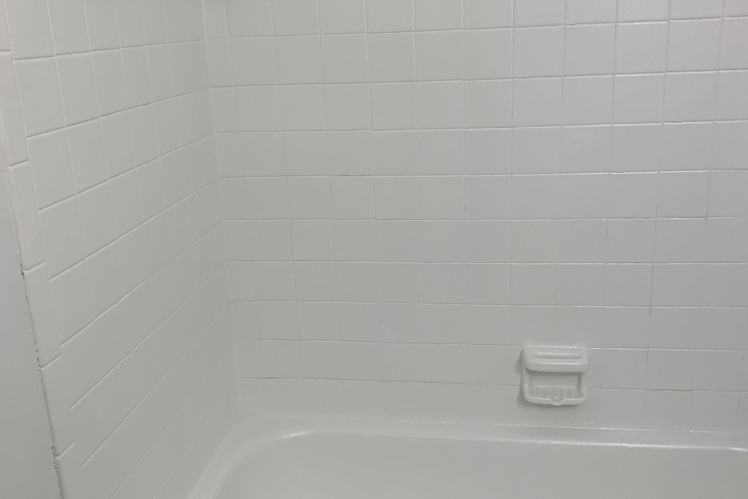 A clean, white tiled bathroom shower stall with a smooth tub surface. The walls are lined with square white tiles, and a small white soap dish is mounted on the wall. The overall appearance is bright and minimalistic, creating a simple and uncluttered space.