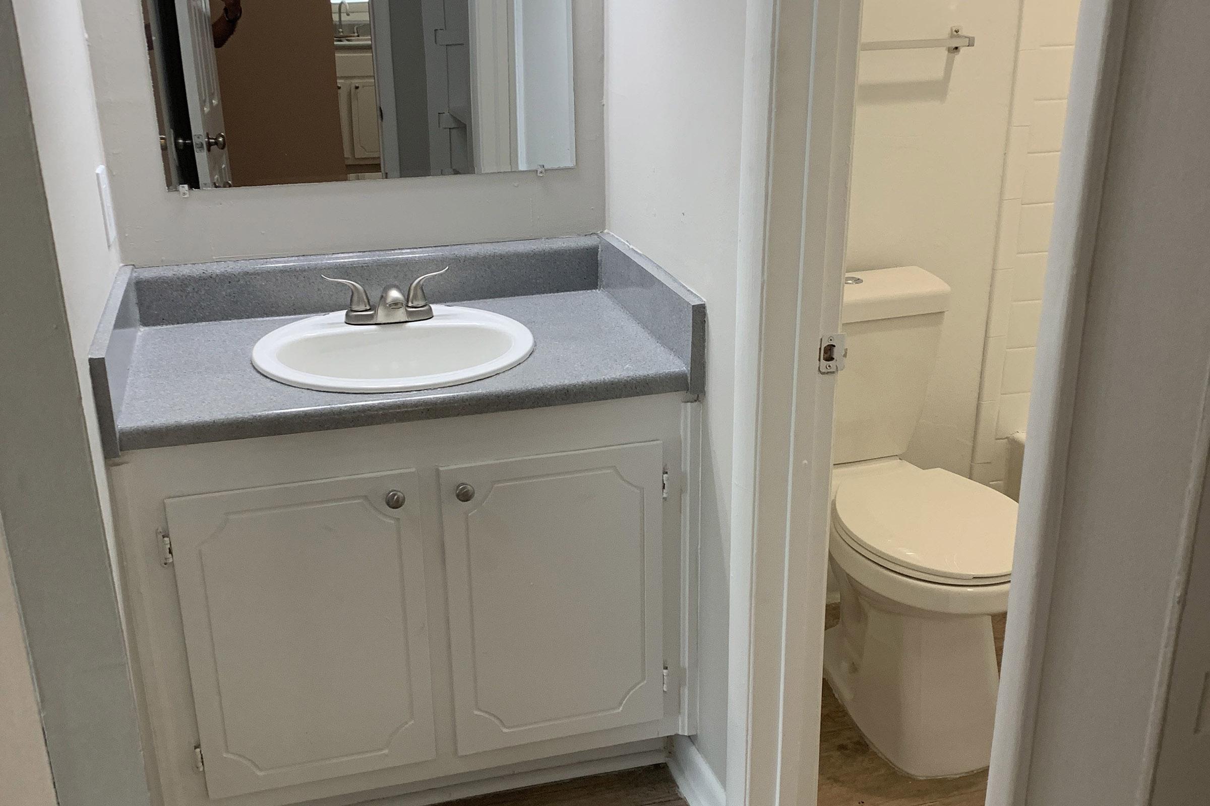 A small bathroom featuring a white vanity with a sink and a mirror above it. Next to the vanity, there is a modern toilet. The walls are painted white, and the flooring is a light-colored wood or laminate. The space appears clean and well-maintained.