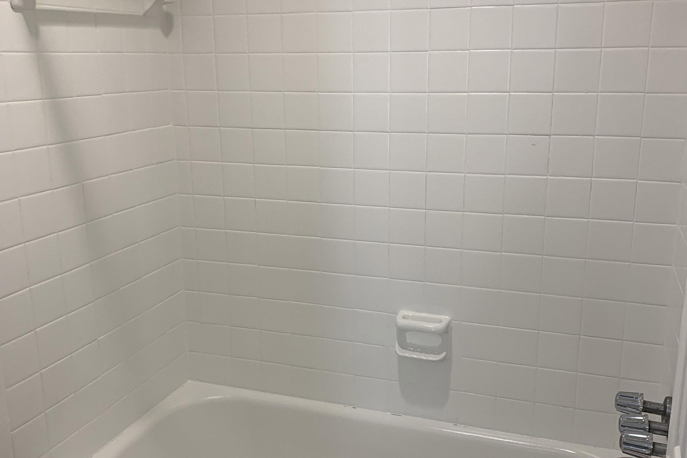 Clean, white tiled bathroom corner featuring a bathtub and a wall-mounted soap dish. The space is well-lit with no visible fixtures or decor, providing a minimalistic and modern appearance.
