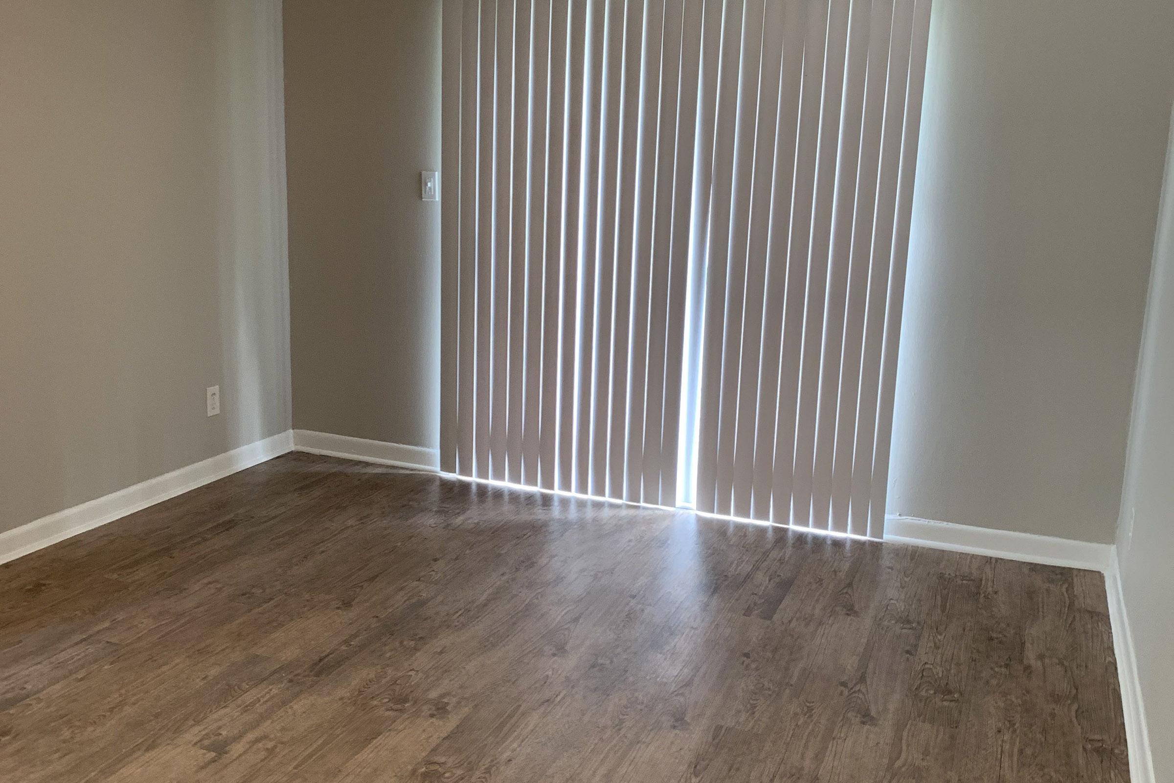 A spacious, empty room with light brown wooden flooring. The walls are painted in a soft neutral color. A vertical blind window allows natural light to stream in, casting shadows on the floor. The room appears freshly cleaned and ready for furnishings.
