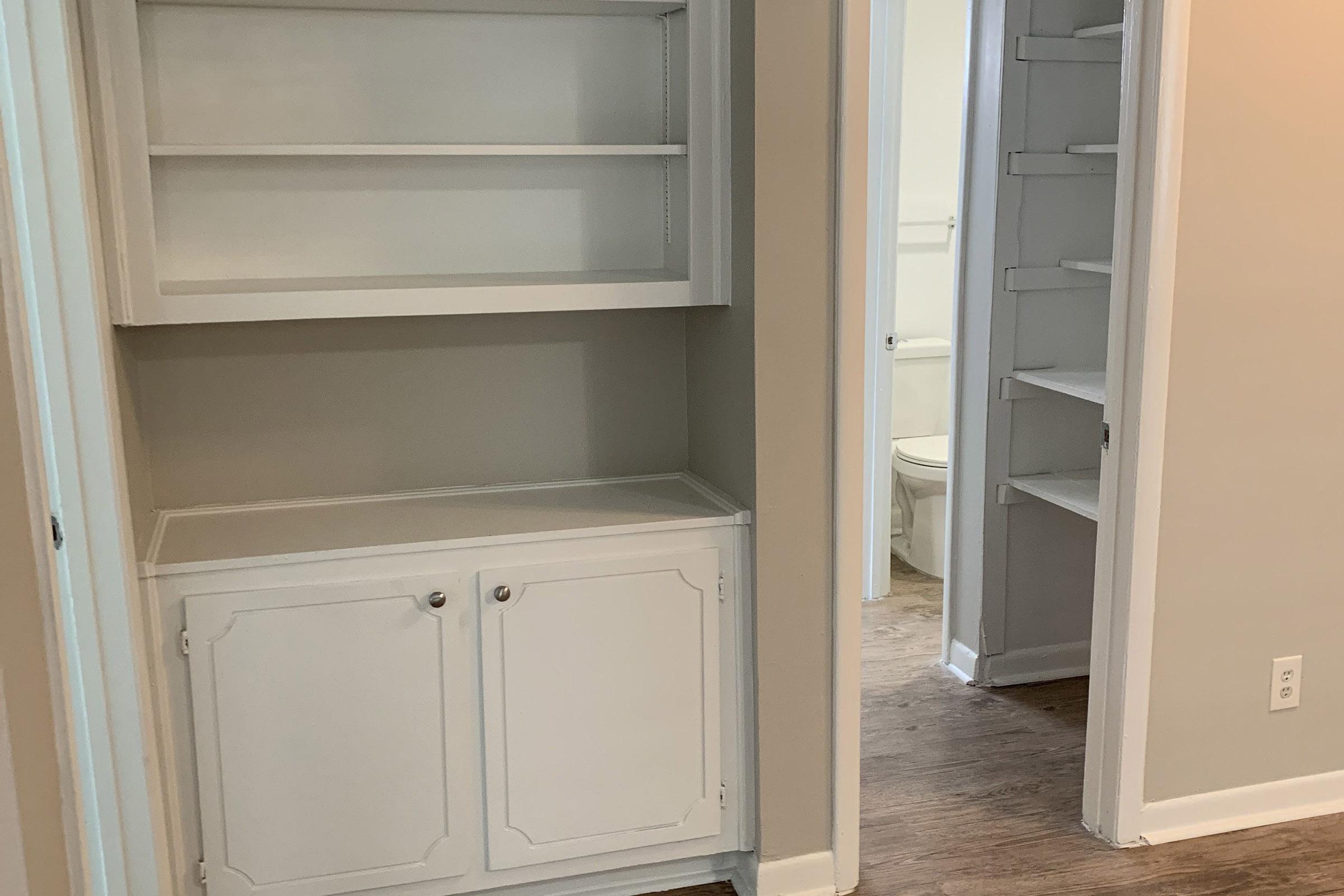 A view of a hallway featuring a white built-in cabinet on the left with open shelves above and closed storage below. On the right, a narrow closet with shelving can be seen. The walls are a light beige color, and there is a glimpse of a bathroom in the background. The flooring is a light-colored wood laminate.
