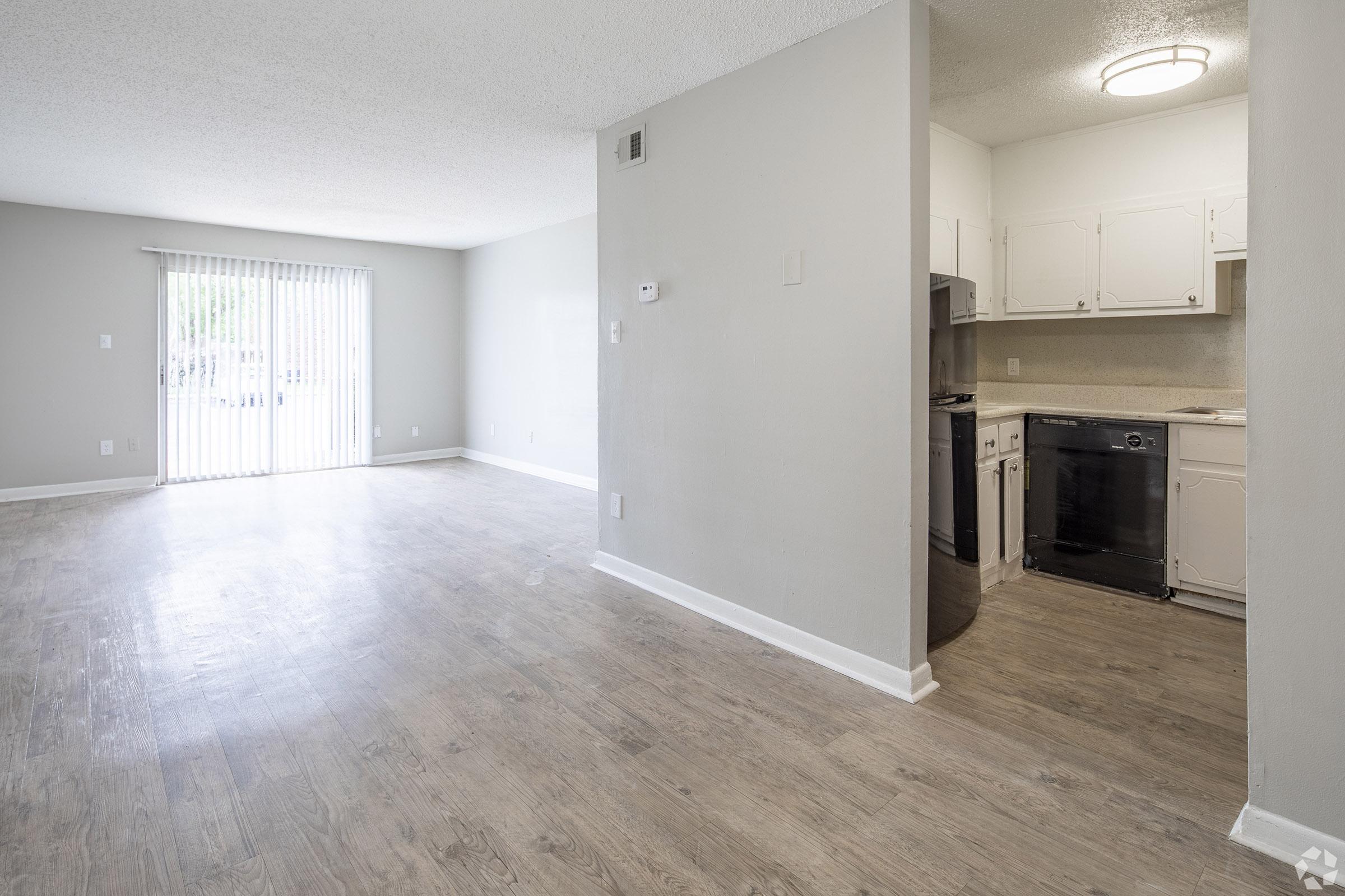 A bright, open living space featuring light gray walls and a large window with vertical blinds. The area has a wood-like floor. A kitchenette is visible, showcasing white cabinets and appliances, including a dishwasher. The room is clean and spacious, with no furniture present.
