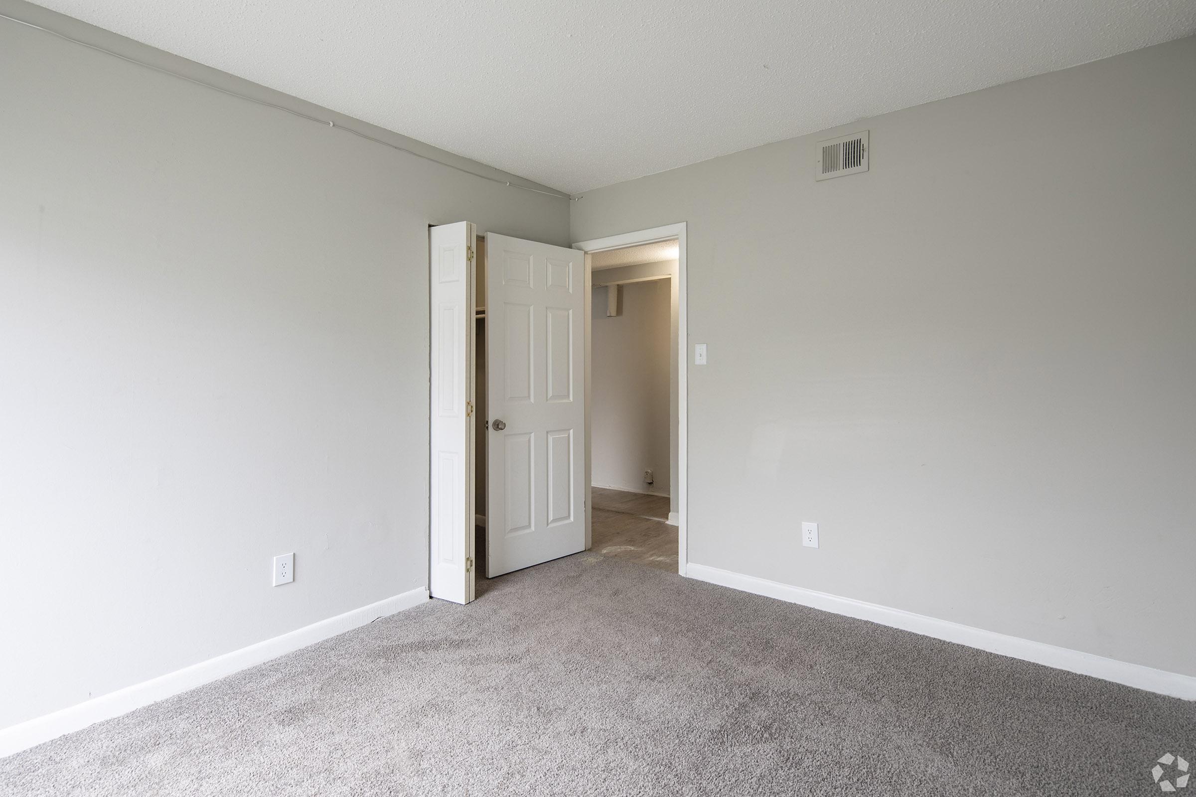 An empty room with light gray walls and carpet, featuring an open door on the left leading to a closet. The space is well-lit with no furniture, showcasing a clean and minimalist design.