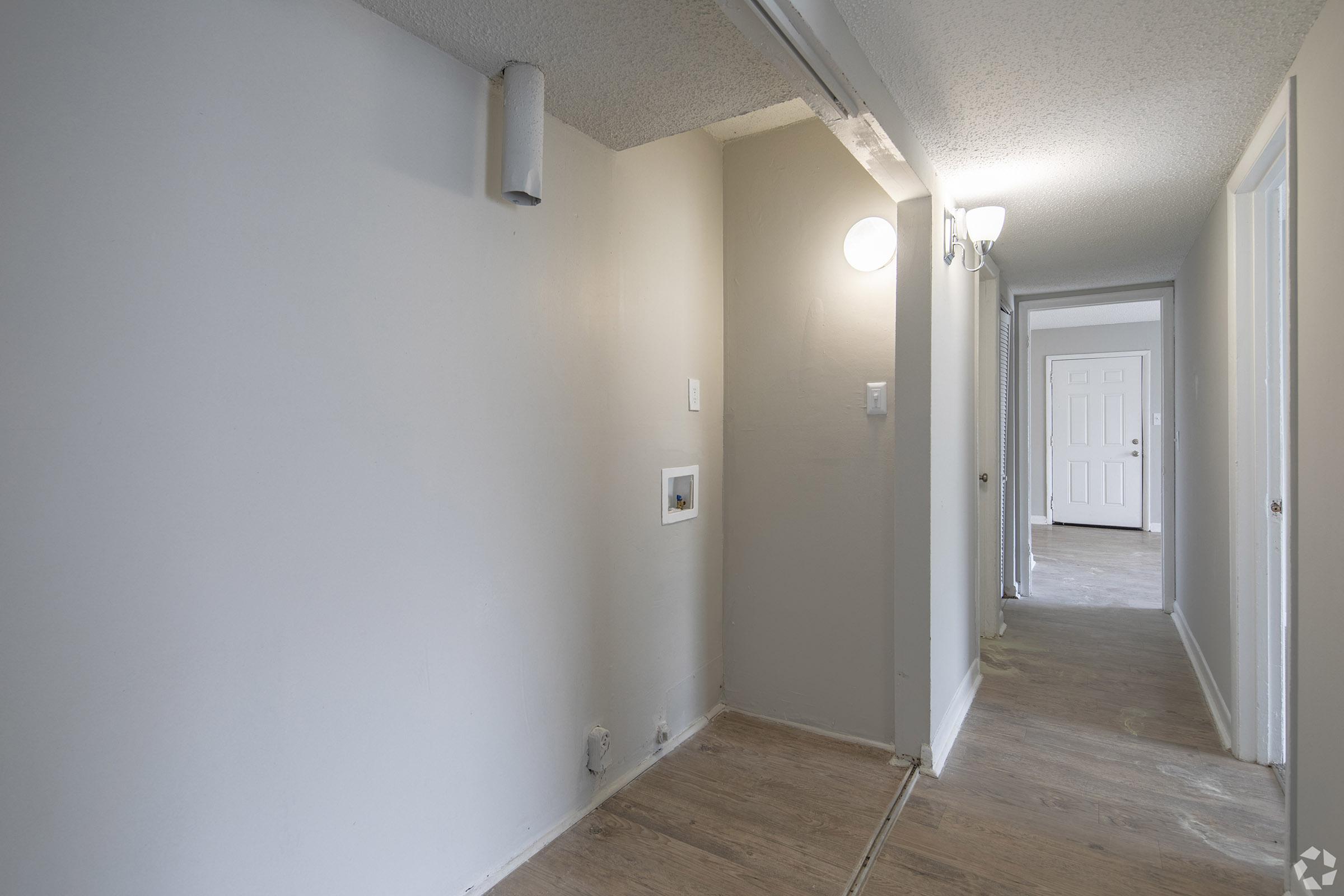 A narrow hallway featuring light-colored walls and a wooden floor. There is a wall-mounted light fixture illuminating the space. On the left side, a small opening is visible, while the corridor leads to a door at the end. The area appears freshly painted and uncluttered.
