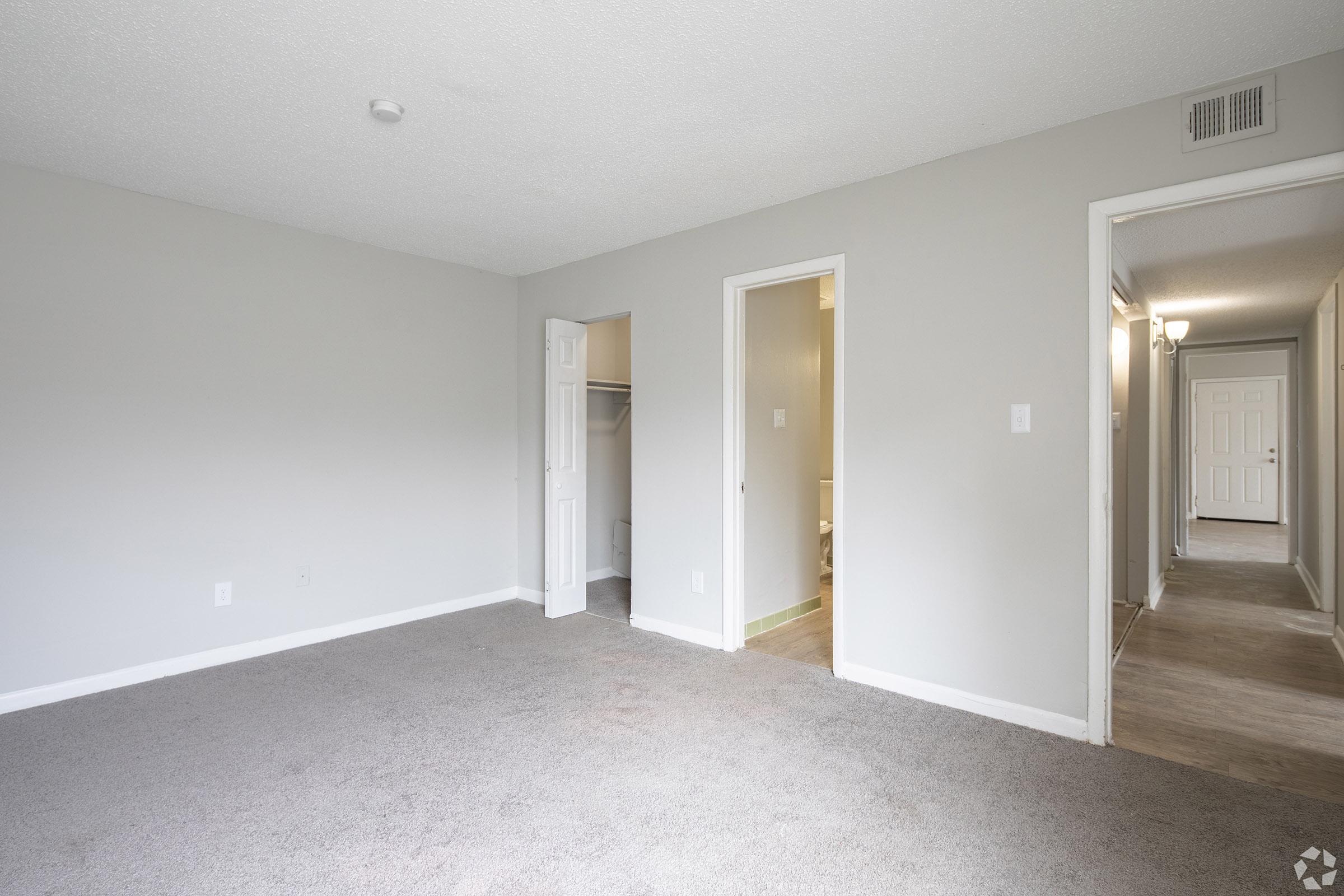 A spacious, light gray room with a carpeted floor, featuring a partially opened closet door on the left and two entrances to other rooms. The walls are painted light gray, and there is a small ceiling light. A hallway can be seen in the background leading to additional rooms.
