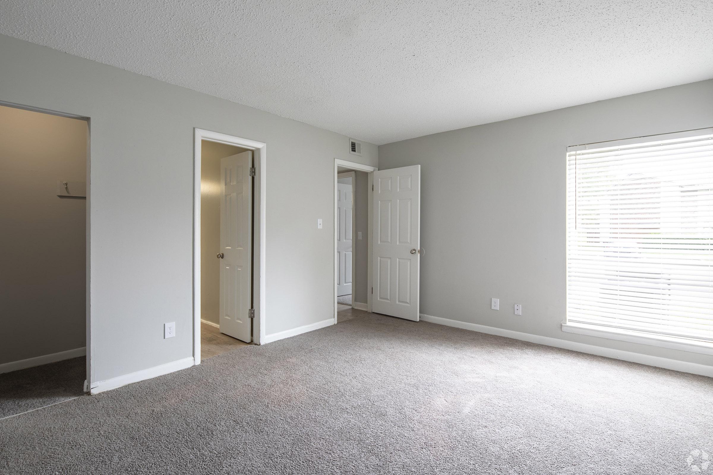 A vacant room with light gray walls and carpeted flooring. There are three open doors leading to other spaces, including one to a closet. A window with blinds lets in natural light, illuminating the room's simple, neutral decor.