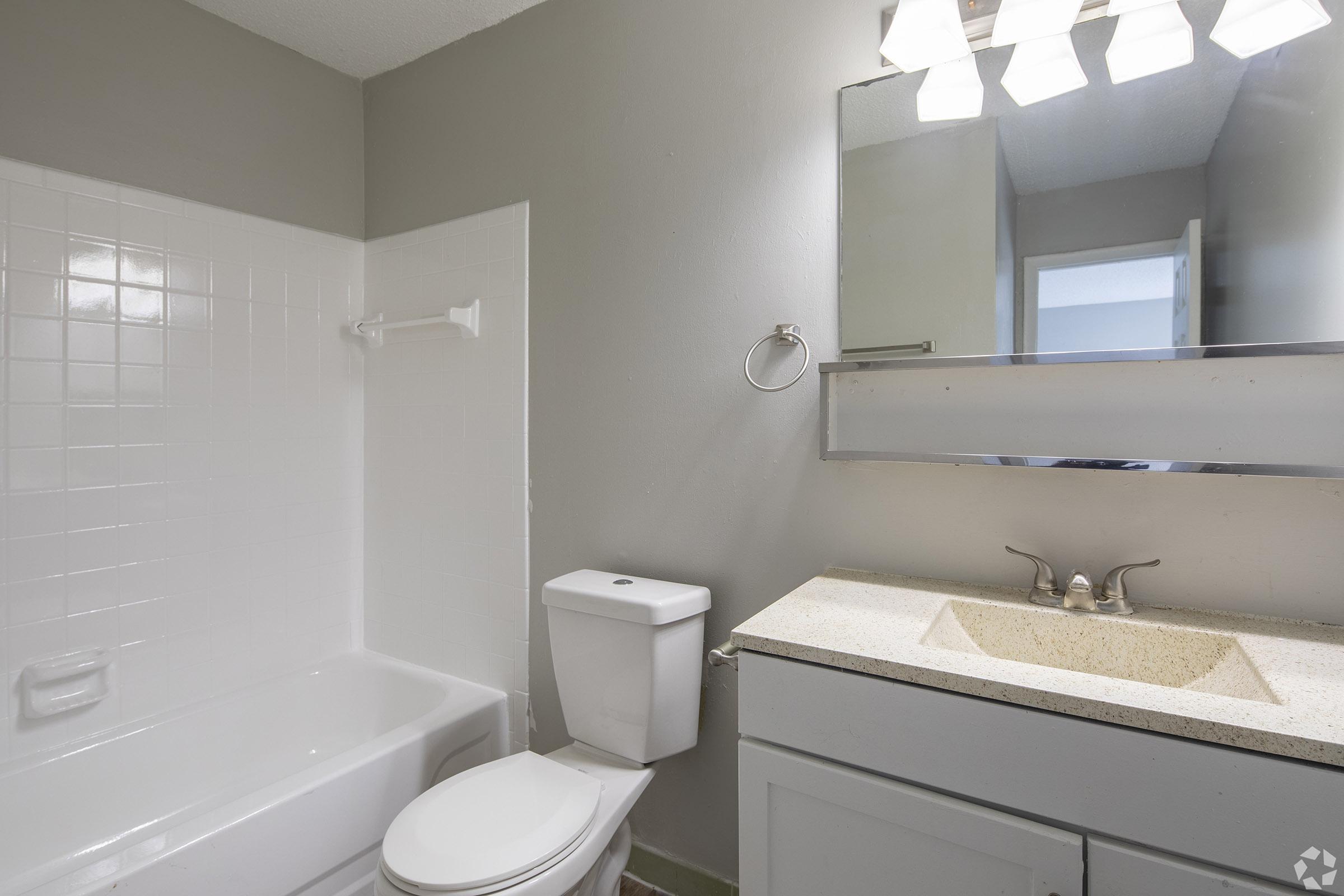 A clean, modern bathroom featuring a white bathtub with a shower, a toilet, and a vanity with a light-colored countertop. There is a large mirror above the sink, and the walls are painted in a soft gray tone. The overall look is spacious and well-lit.