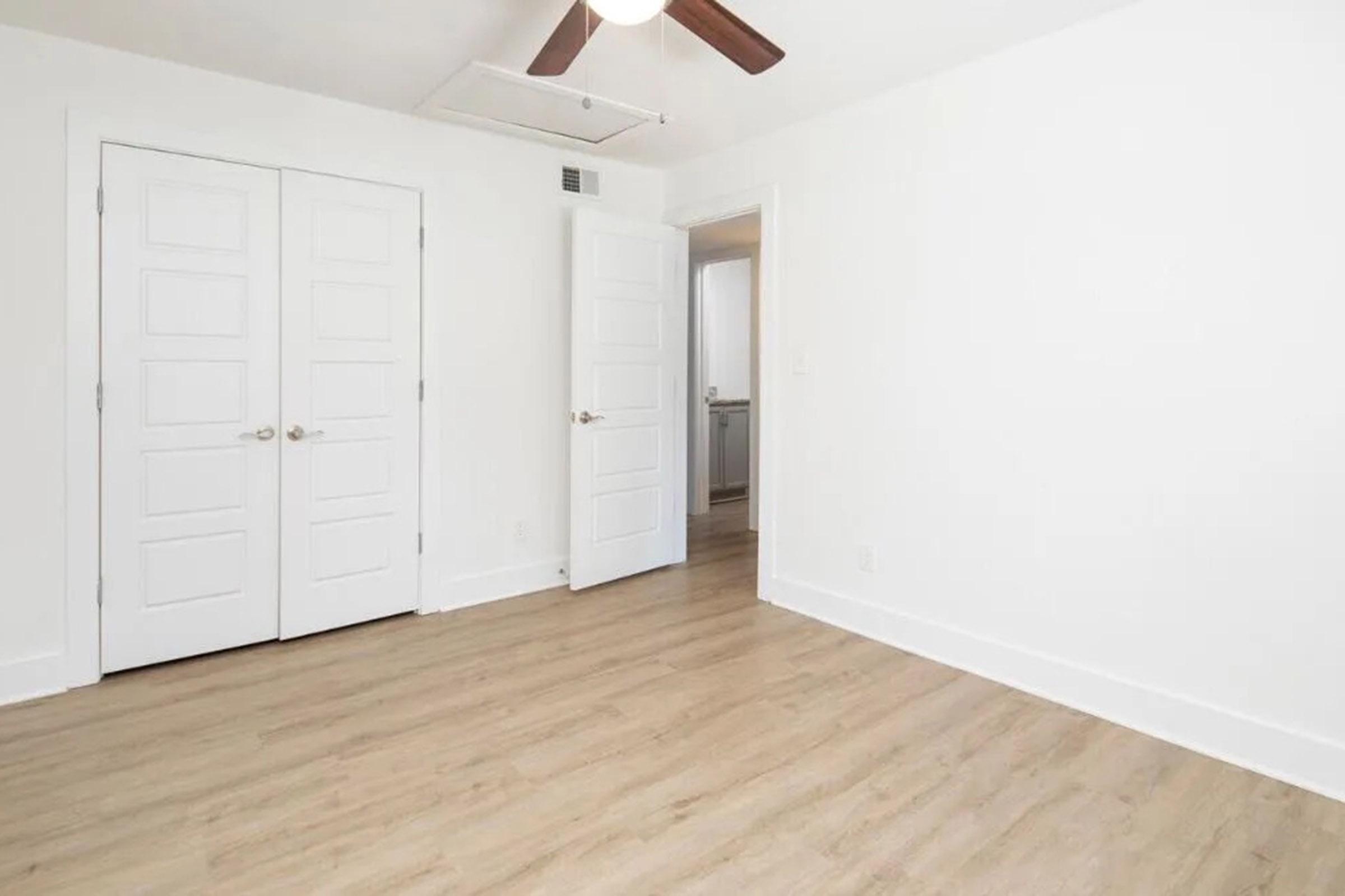 A bright, empty room with light-colored walls and wooden flooring. There are two closed doors with white panels on one wall, and a visible doorway leading to another room in the background. A ceiling fan is mounted above, adding a modern touch to the minimalist space.