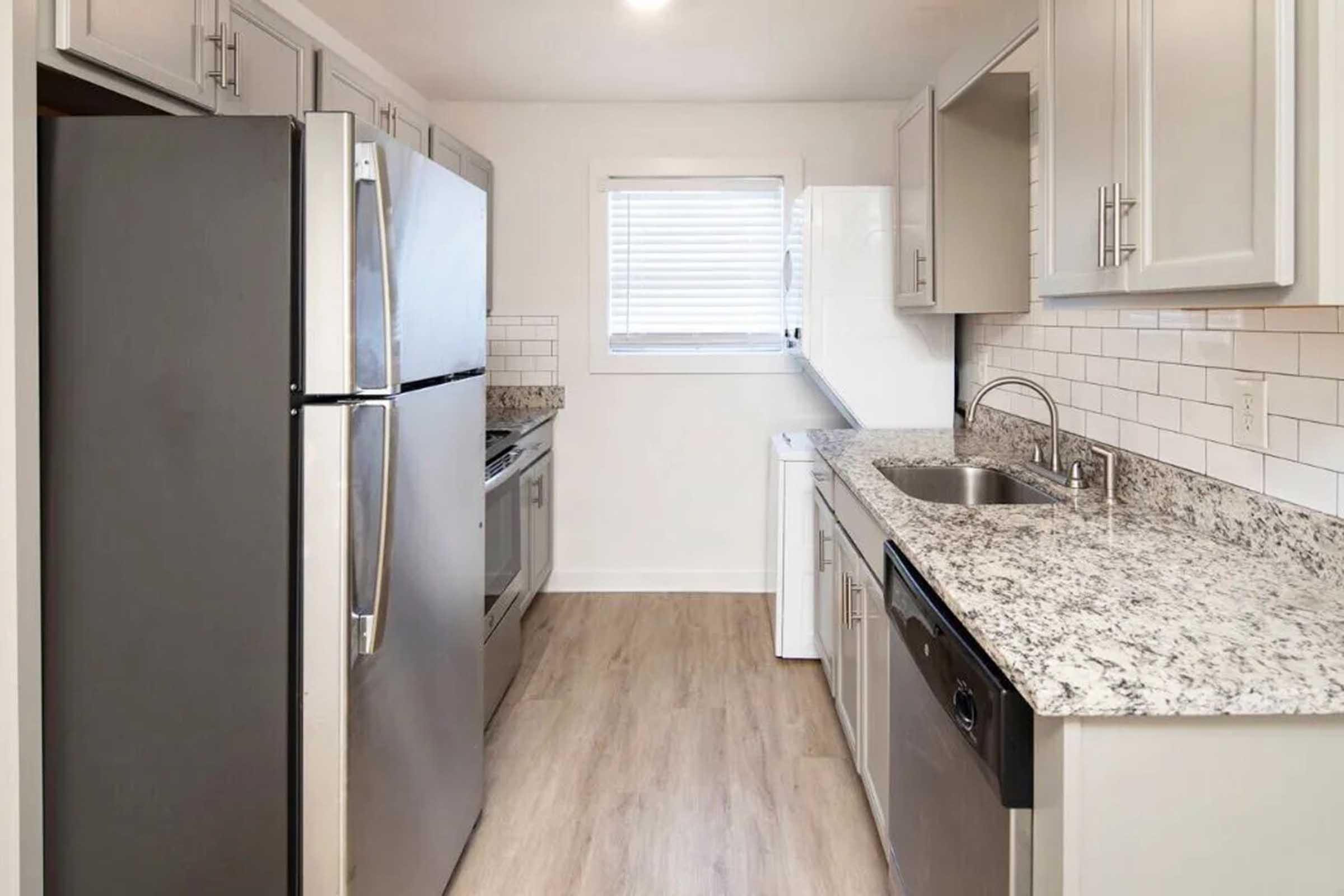 Modern kitchen featuring stainless steel appliances, including a refrigerator, oven, and dishwasher. The countertops are made of granite, complemented by light gray cabinetry. A window provides natural light, and the flooring is a light wood finish, creating a clean and spacious atmosphere.