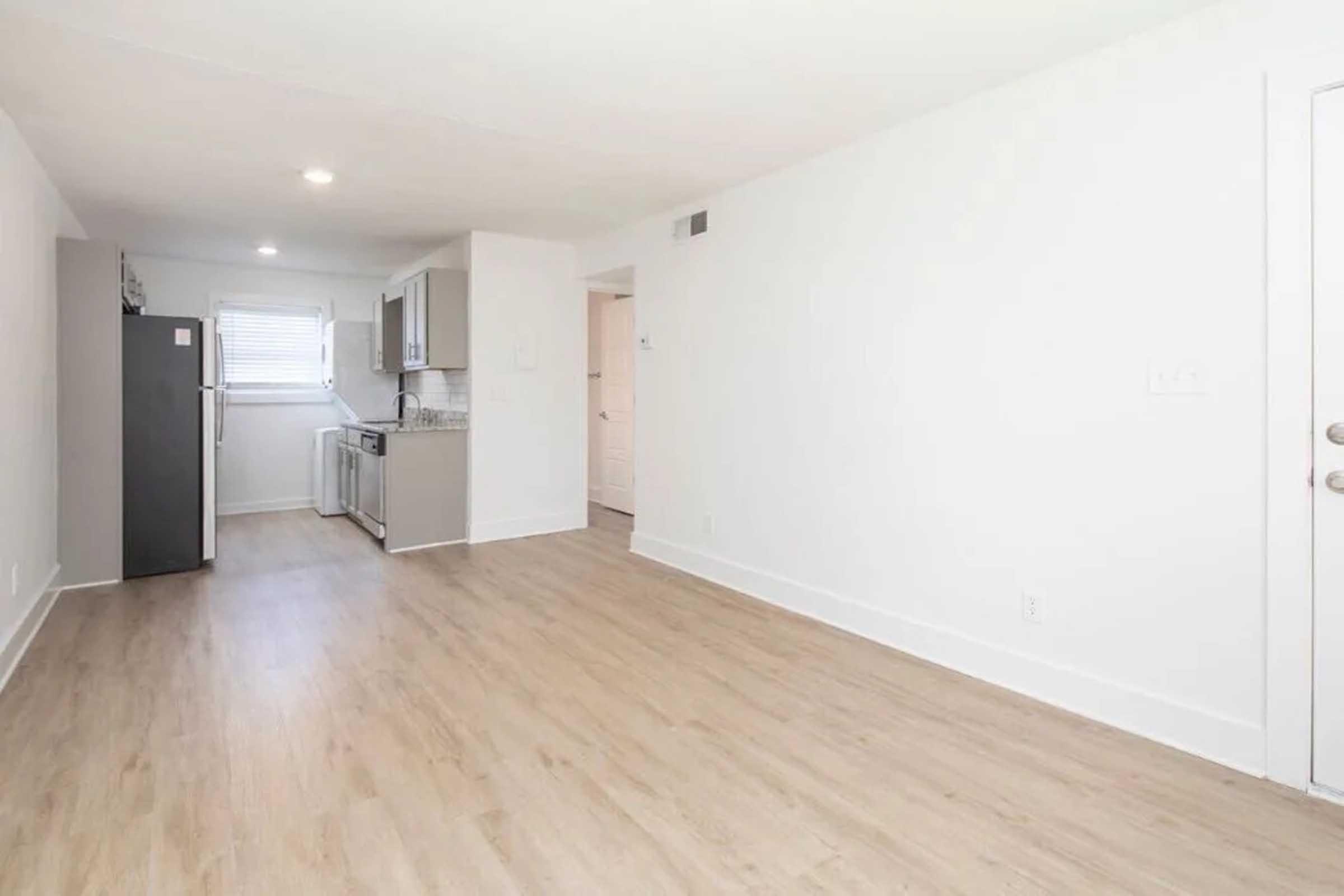 A bright, modern kitchen and living space featuring light wood flooring, neutral-colored walls, and a window allowing natural light. The kitchen includes gray cabinets, a refrigerator, and a stove. The layout is open and spacious, creating a welcoming atmosphere.